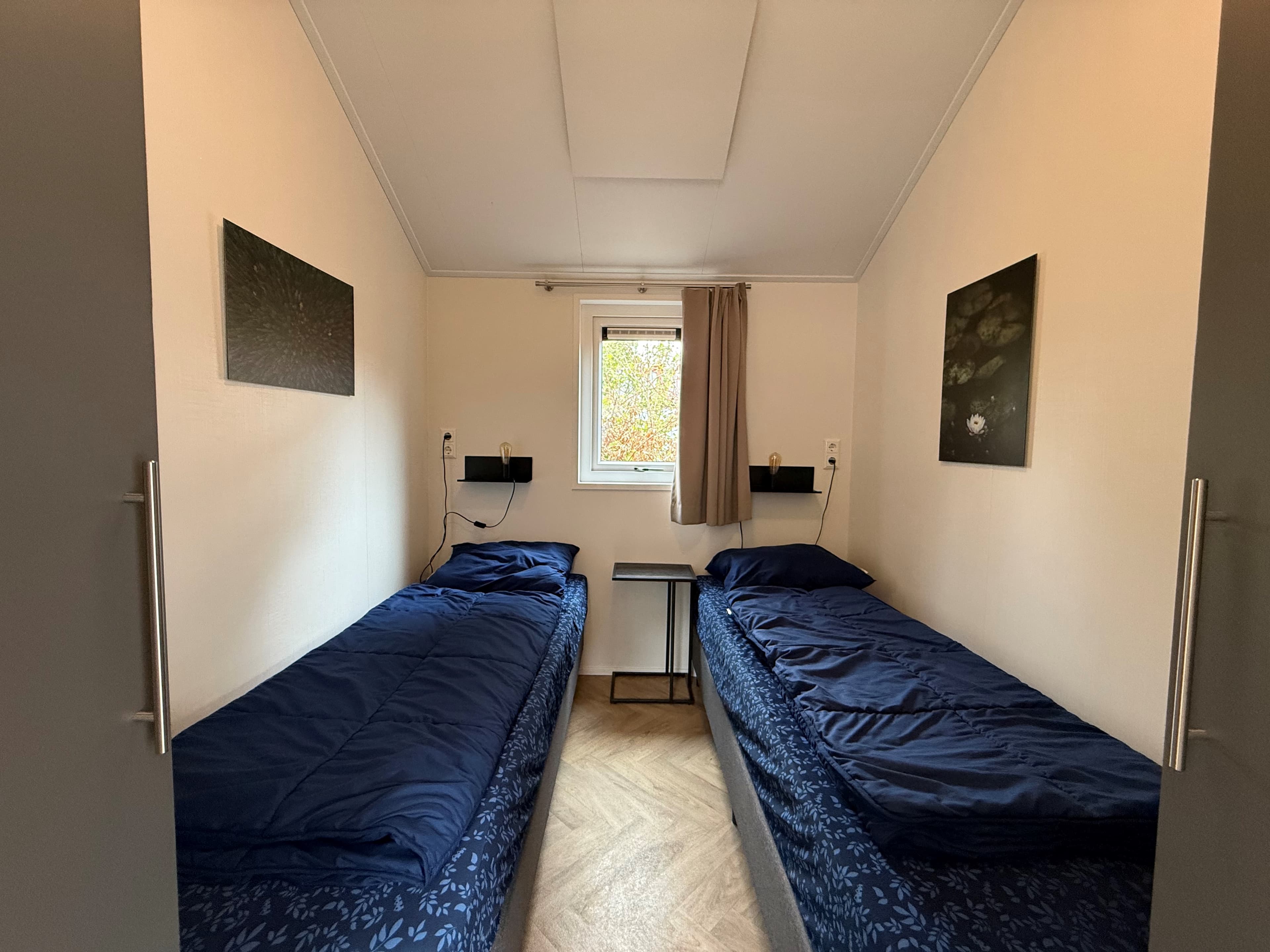 Zijaanzicht van een slaapkamer met twee losse bedden, een klein nachtkastje en een raam met uitzicht op de buitenlucht