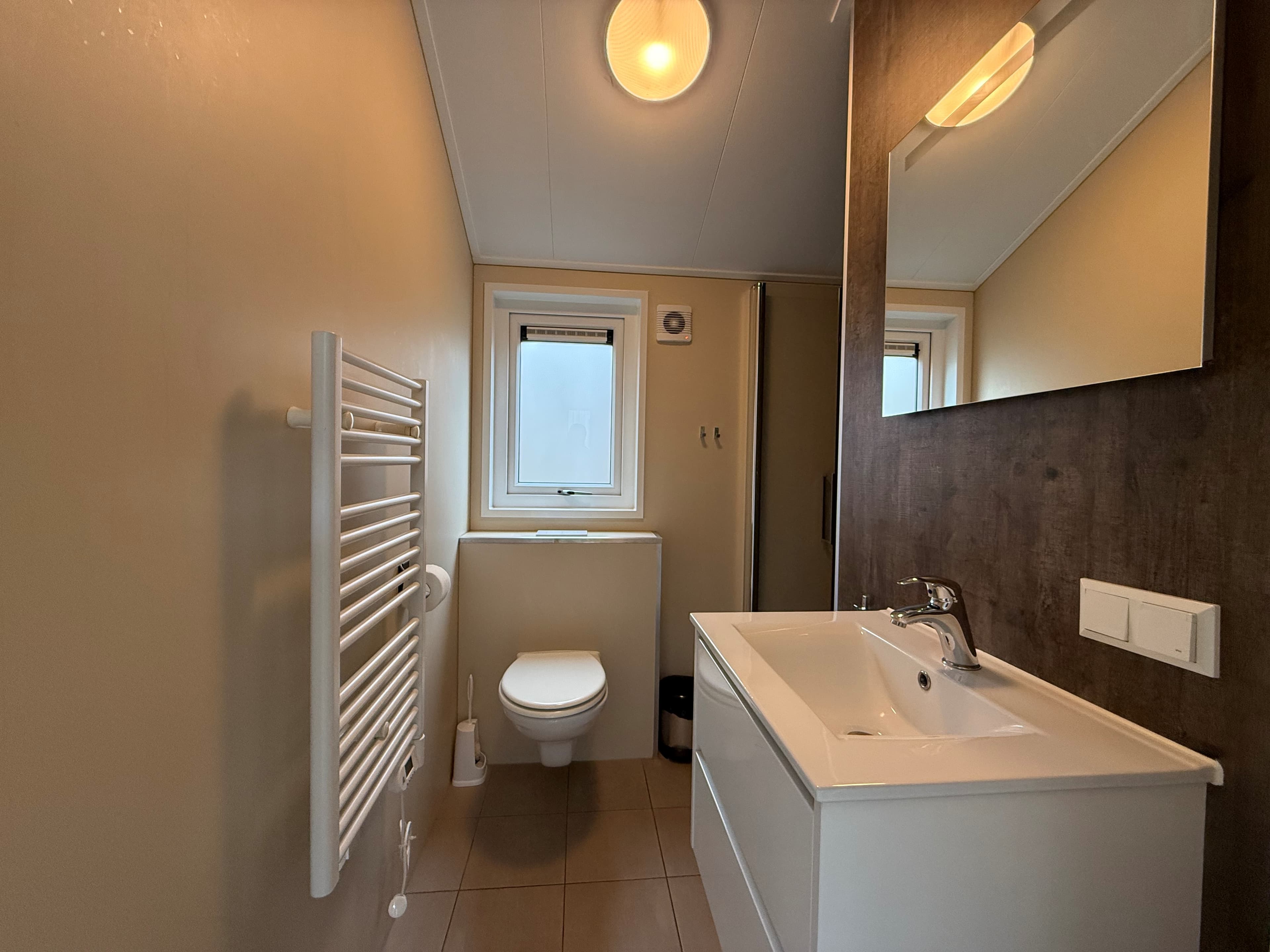 Licht en modern badkamer met toilet, wastafel en spiegel, plus een handdoekenradiator en een raam voor natuurlijk licht