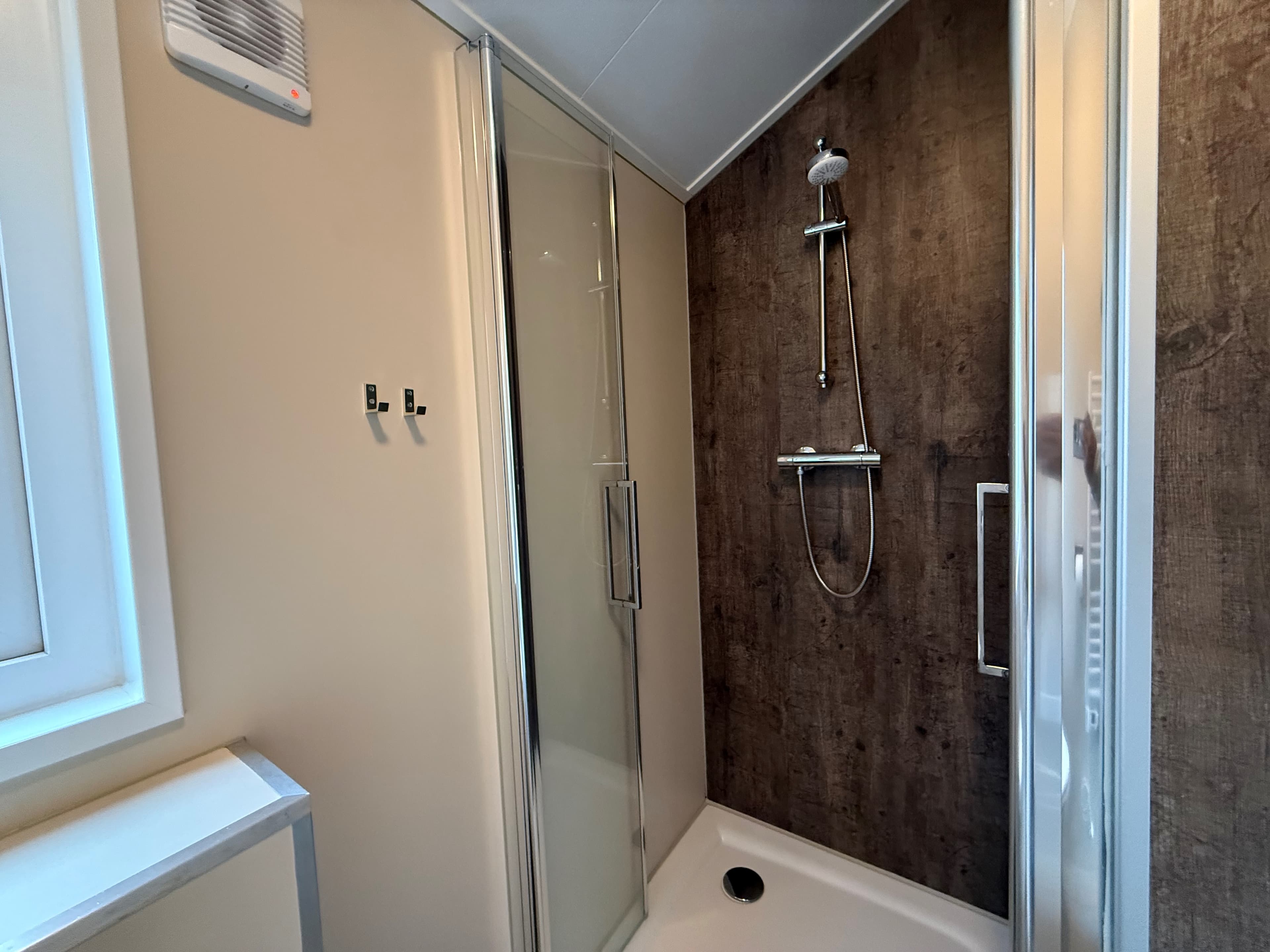 Douchecabine met glazen deur, wandpanelen in houtlook en een regendouchekop. Plafond met verlichting.