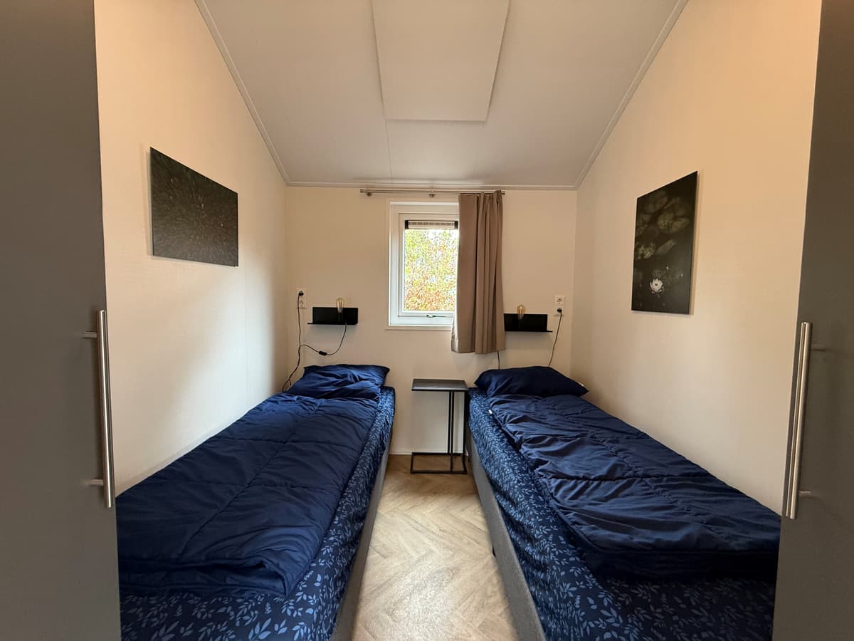 Zijaanzicht van een slaapkamer met twee losse bedden, een klein nachtkastje en een raam met uitzicht op de buitenlucht