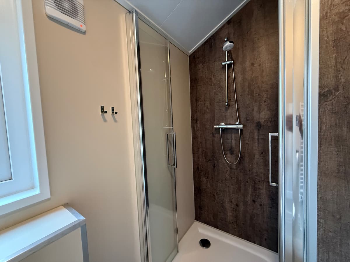 Douchecabine met glazen deur, wandpanelen in houtlook en een regendouchekop. Plafond met verlichting.