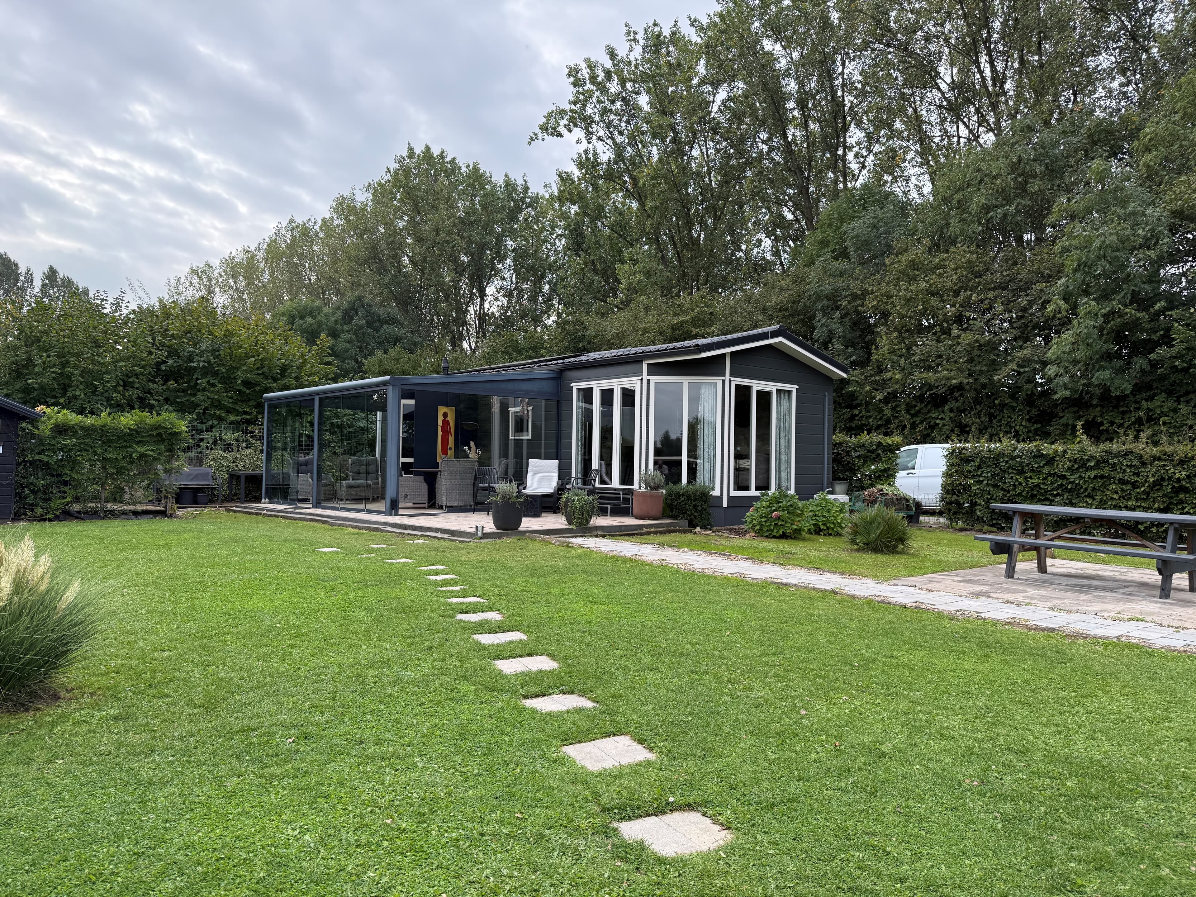 Recreatiewoning met een beschutte veranda, groene grasveld en een pad van stenen, omringd door bomen.
