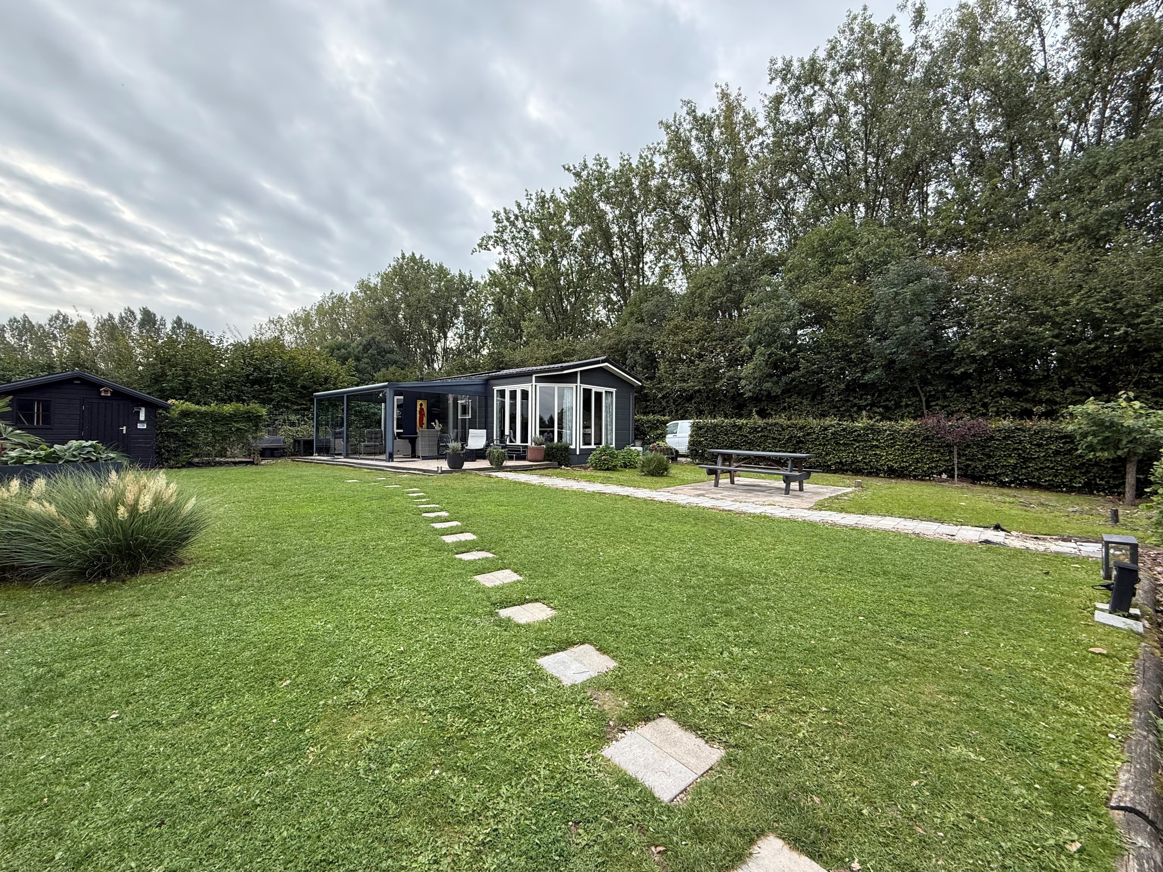 Recreatiewoning met een ruime tuin, grasveld en betegelde paden, omringd door bomen en struiken.