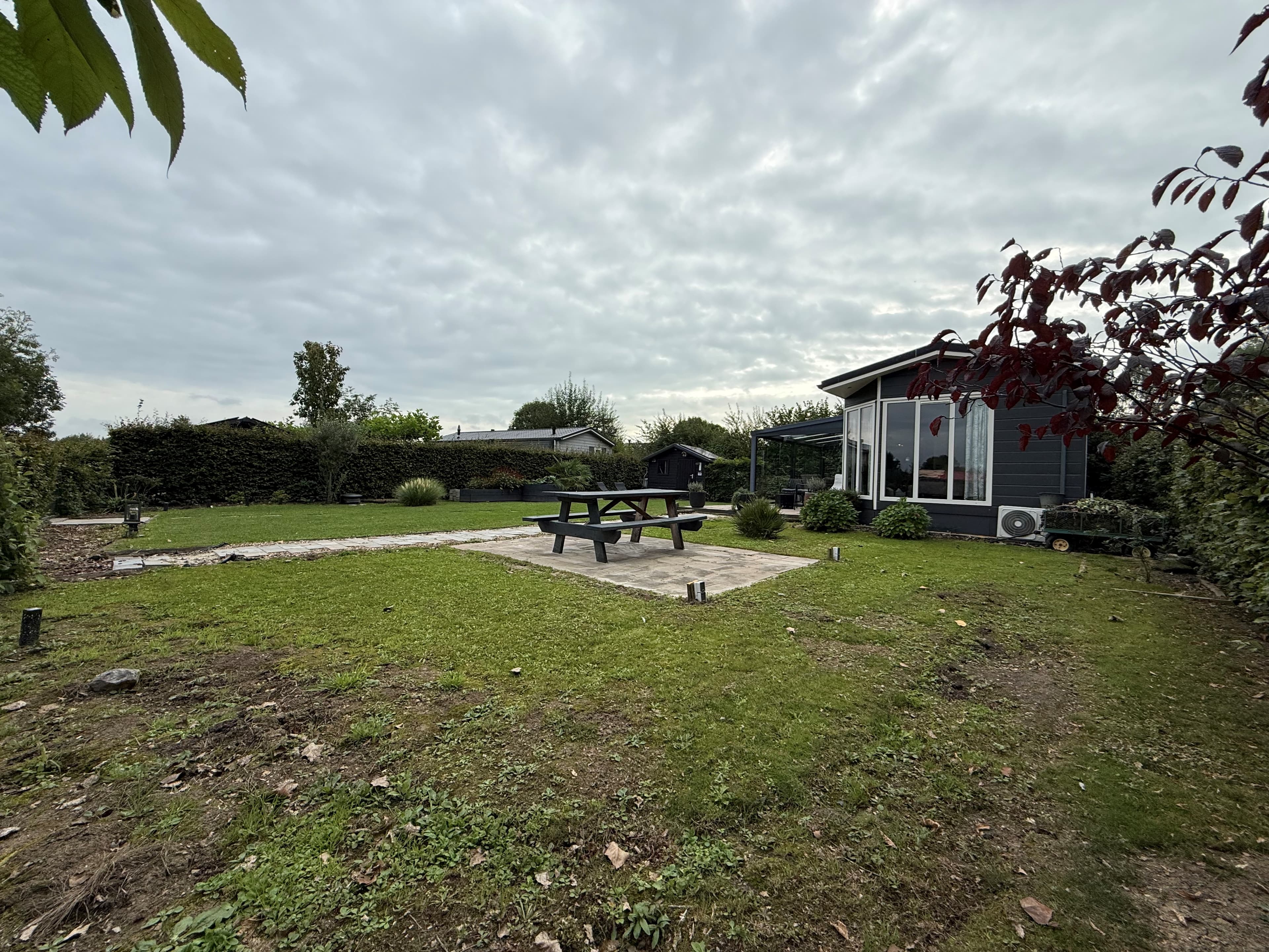 Ruim grasveld met een picknicktafel en enkele planten rondom een recreatiewoning, onder een bewolkte lucht