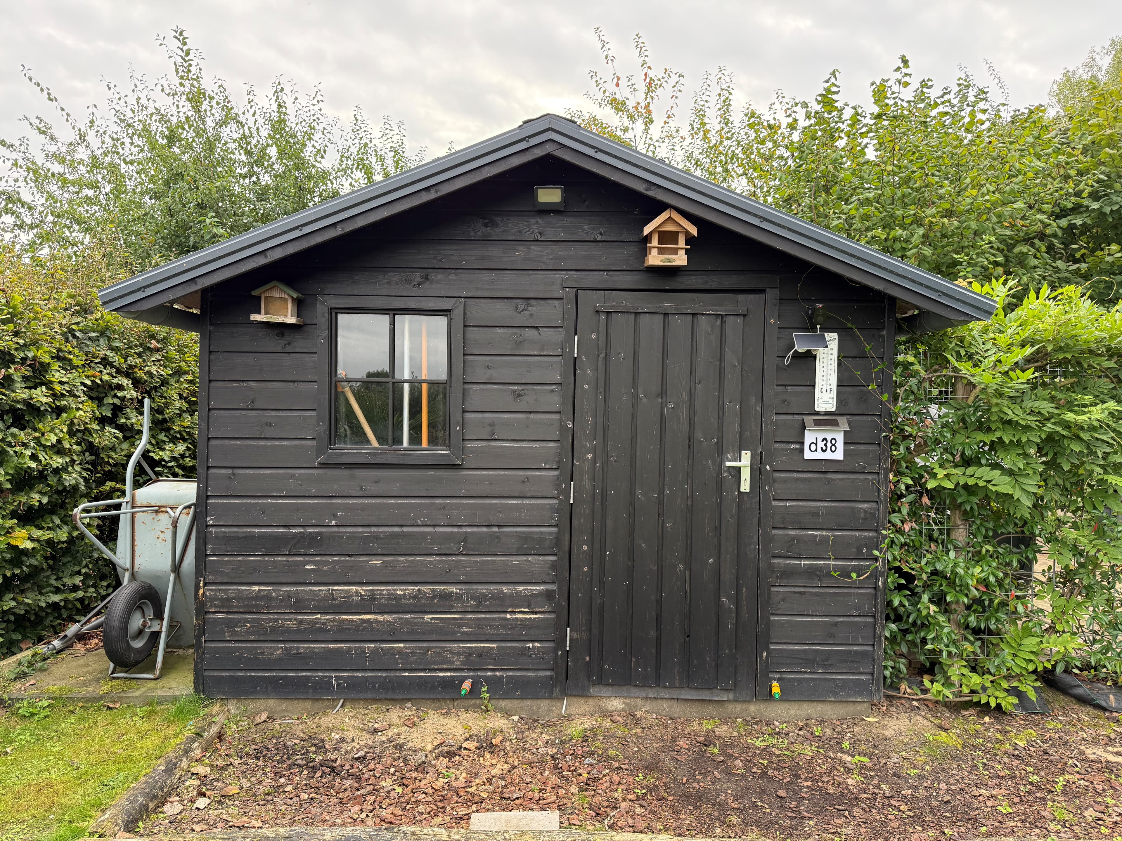 Zwart houten recreatiewoning met een deur en een raam, omgeven door groen en een wheelbarrow aan de zijkant