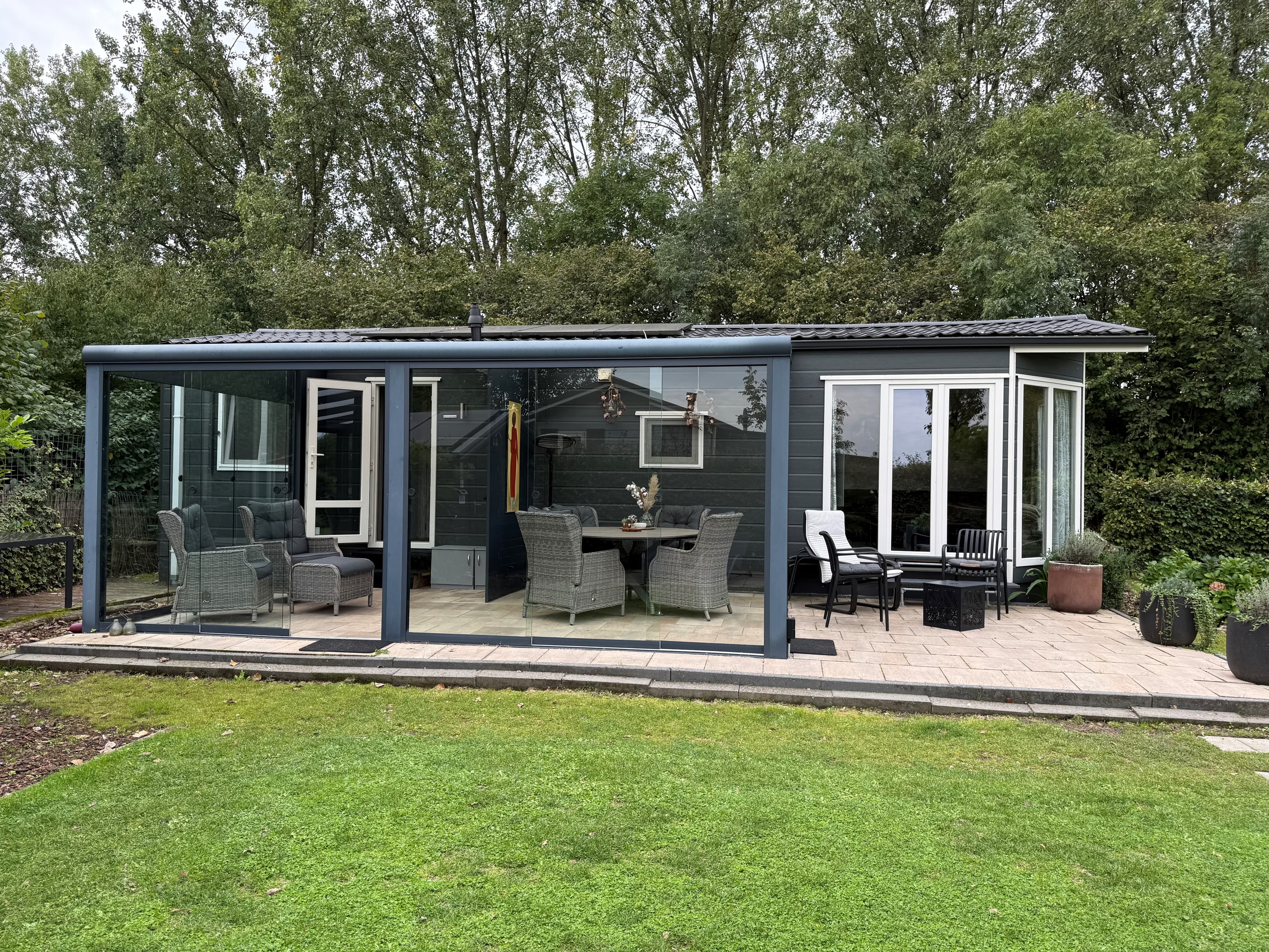 Moderne recreatiewoning met een glazen overkapping en buitenmeubilair in een groene omgeving