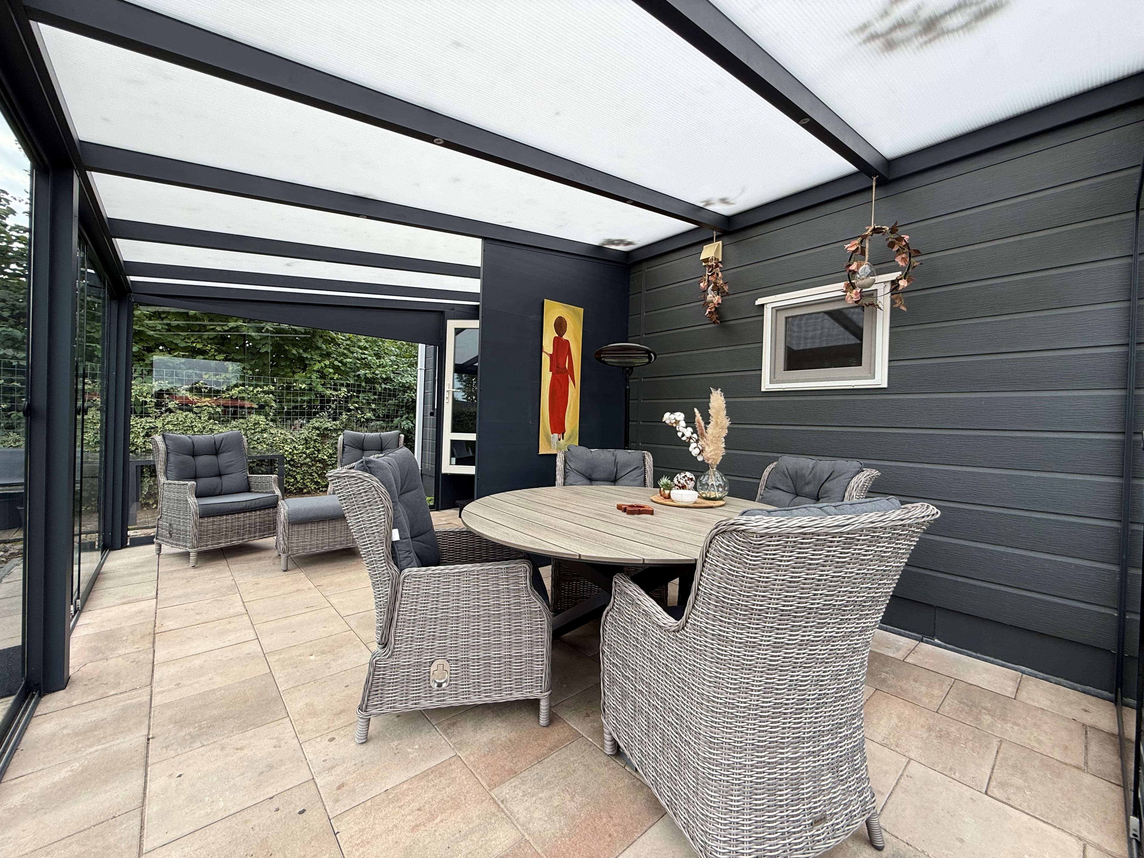 Overdekte patio met een ronde tafel en comfortabele wicker stoelen, omgeven door glazen wanden en groene planten.
