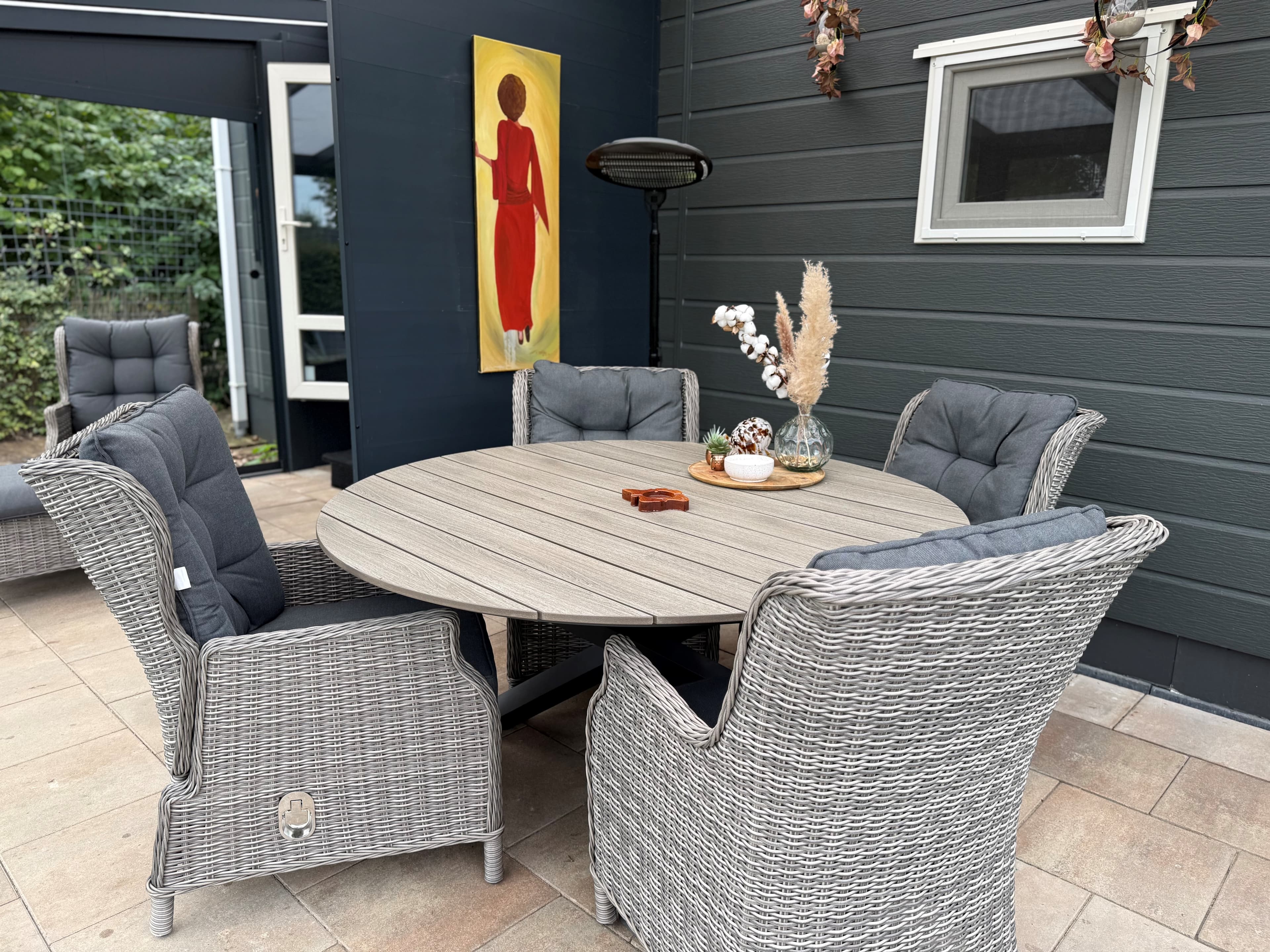 Tuinset met vier grijze stoelen rond een ronde tafel, decoratief schilderij aan de muur en planten in vaas in een overdekte r