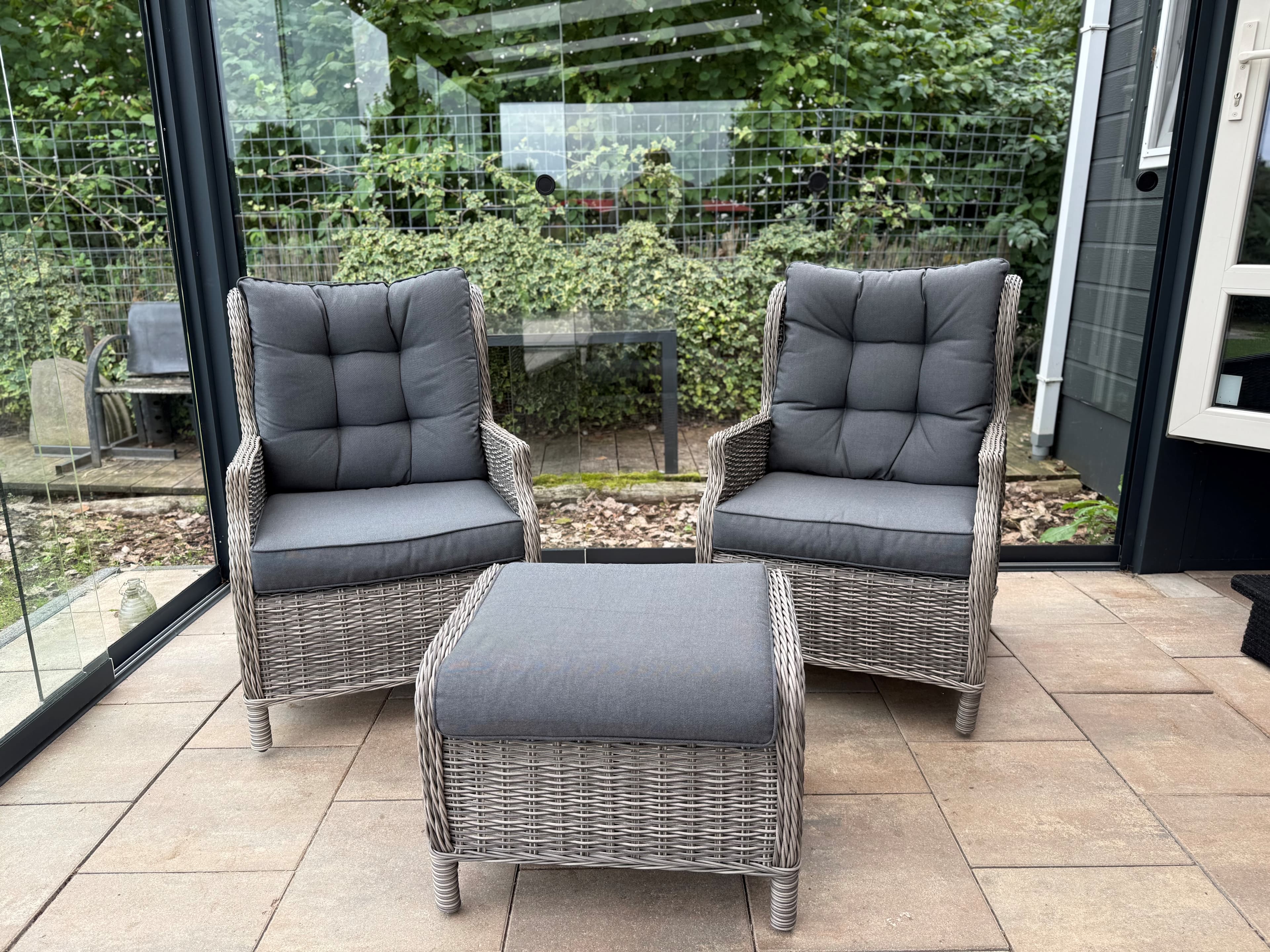 Twee comfortabele wicker stoelen met grijze kussens en een bijpassende tafel in een glazen serre.