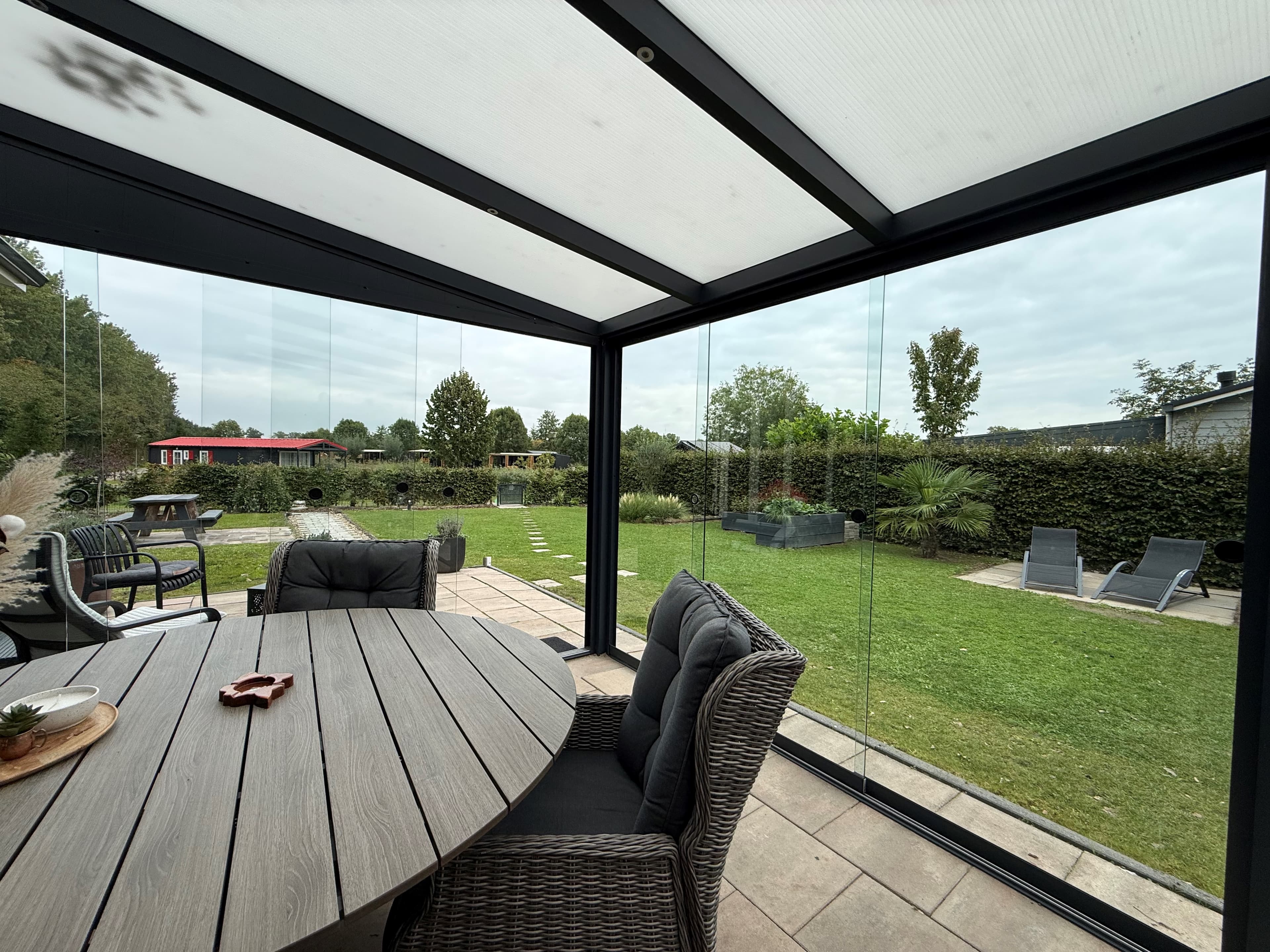 Zicht op een overdekte veranda met een ronde tafel en kuipstoelen, omringd door een groene tuin en loungestoelen.