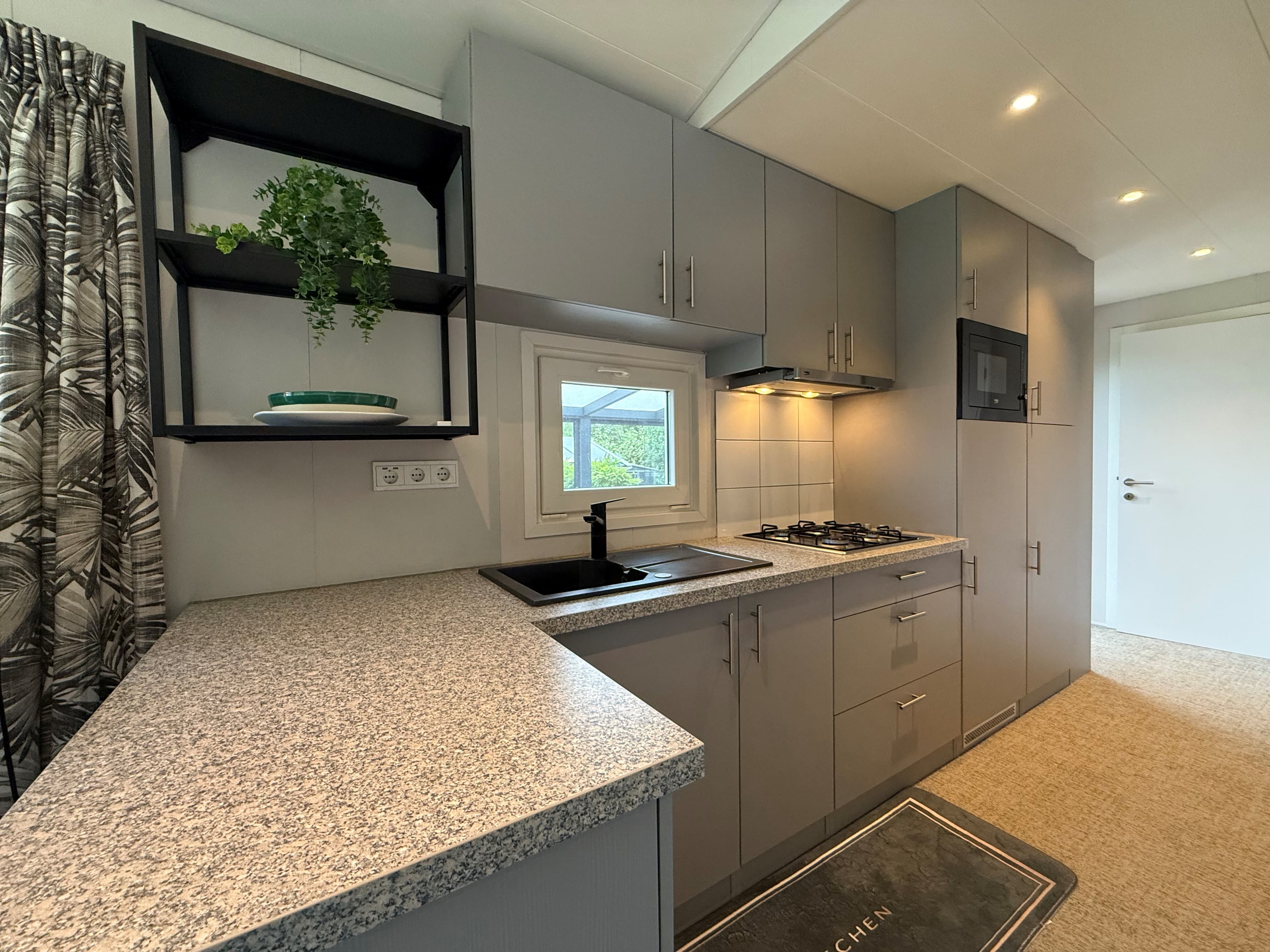 Moderne keuken met grijze kasten, kookplaat en aanrecht, schijnbaar met natuurlijke lichtinval en een plantenhouder