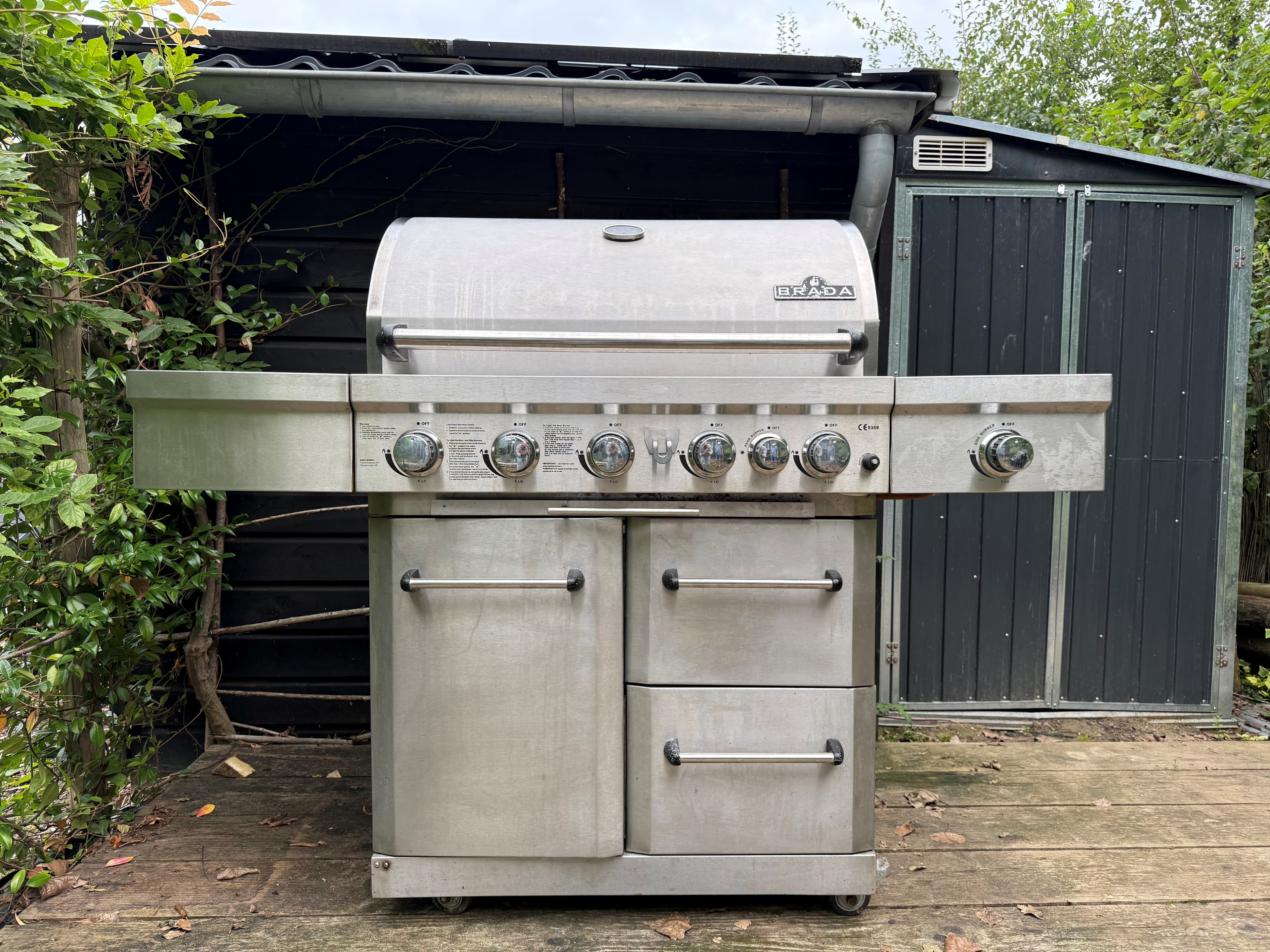 RVS barbecue met meerdere branders op een houten terras, achter een groene achtergrond van bomen en een schuur.