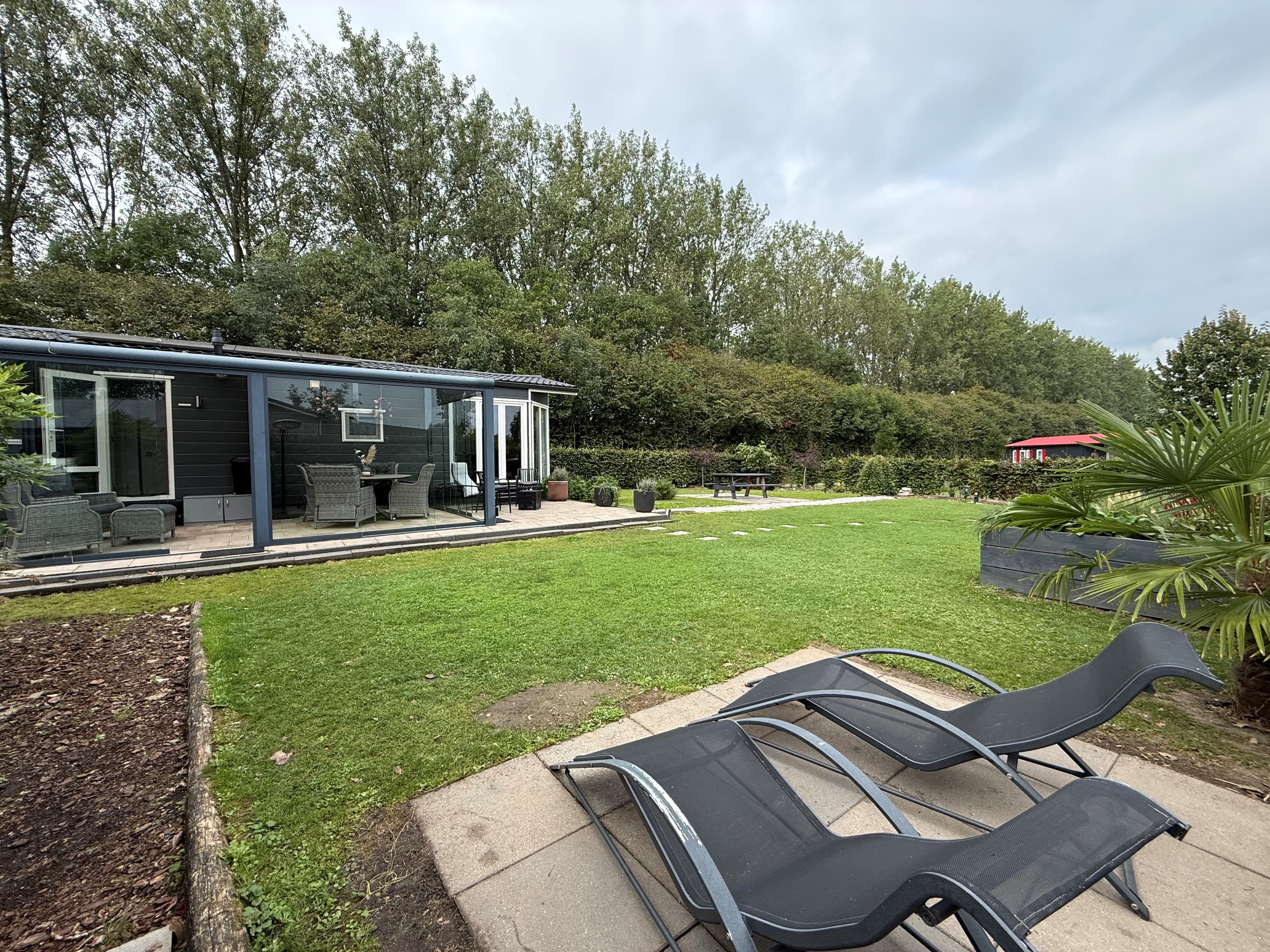 Recreatiewoning met groen gazon, terras en ontspanningsstoelen, omgeven door bomen en struiken.