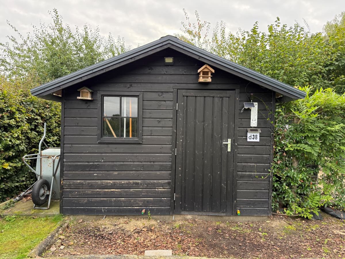 Zwart houten recreatiewoning met een deur en een raam, omgeven door groen en een wheelbarrow aan de zijkant
