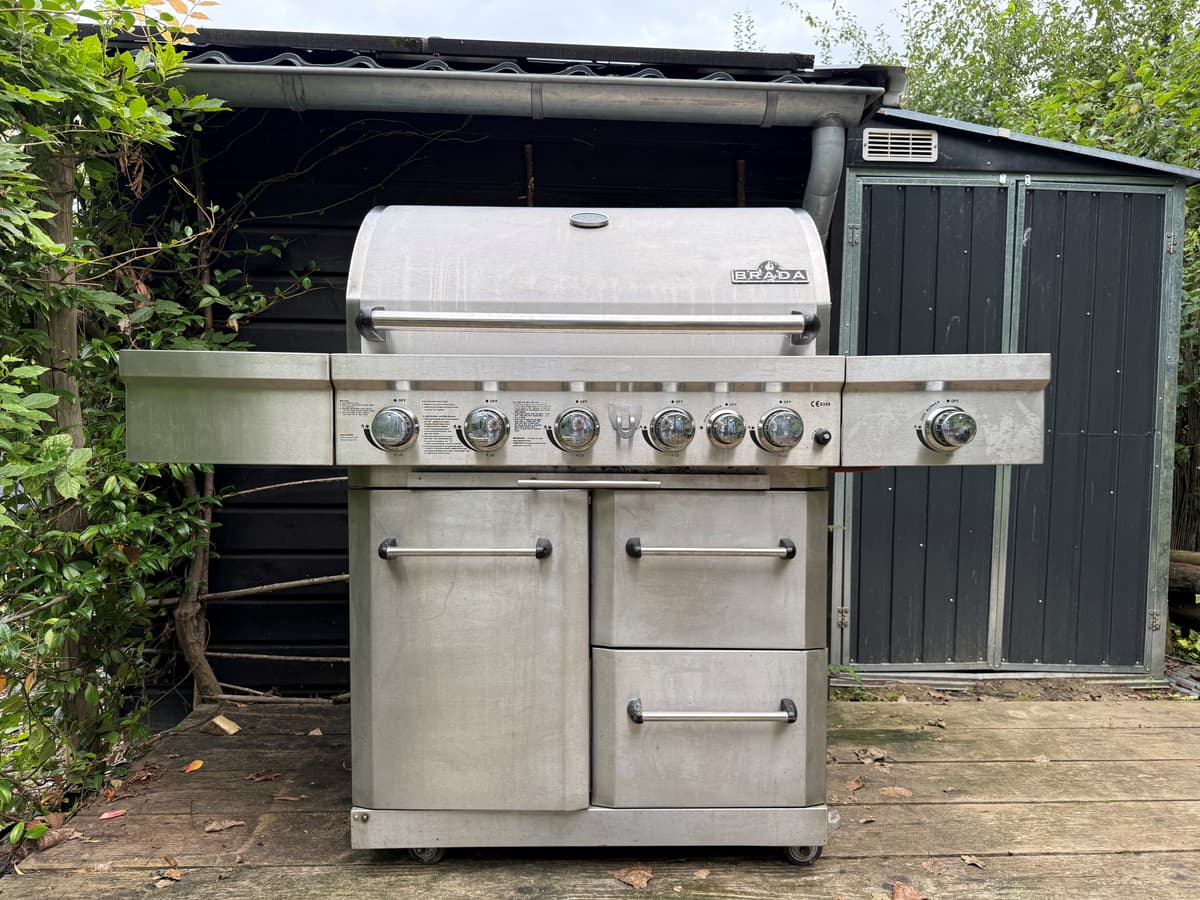 RVS barbecue met meerdere branders op een houten terras, achter een groene achtergrond van bomen en een schuur.