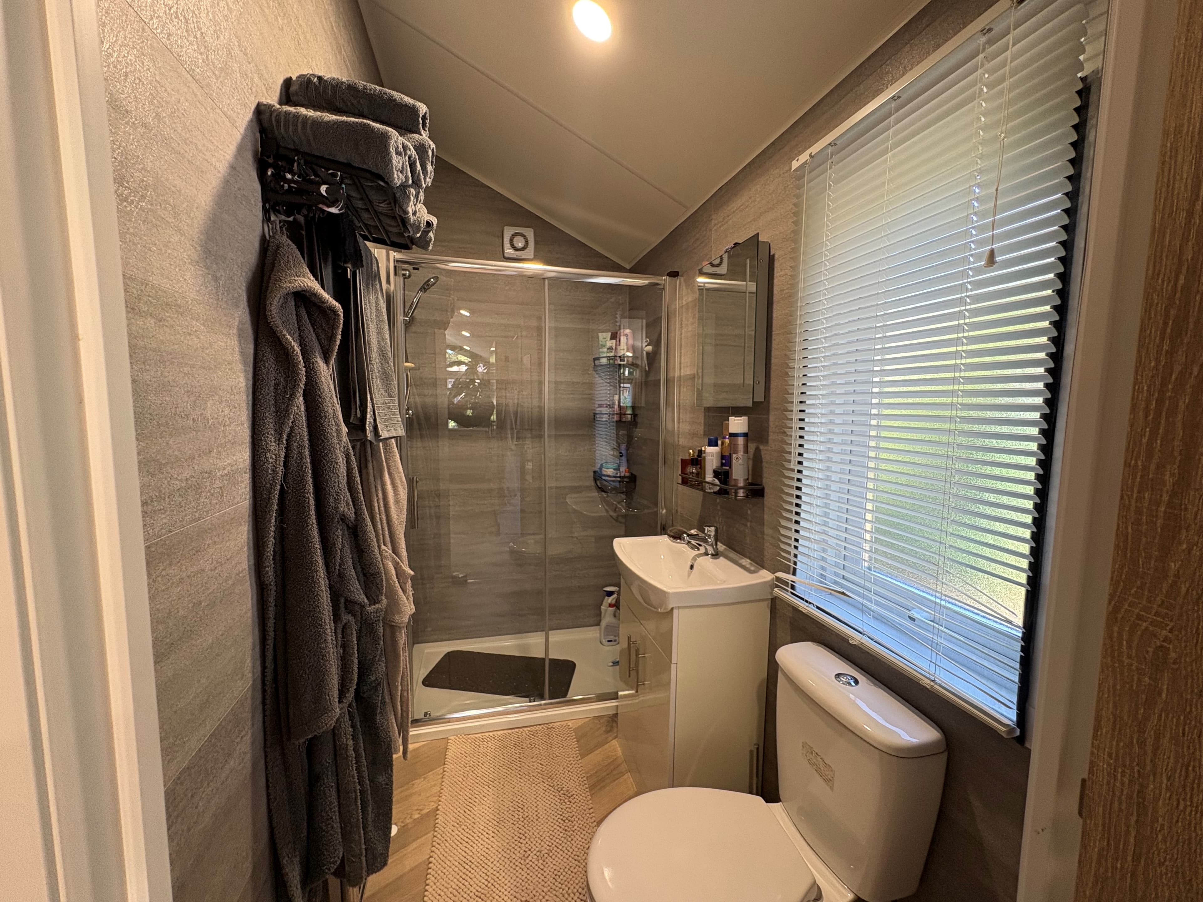 Moderne badkamer met douche, toilet, wastafel en handdoeken; grote ramen met jaloezieën bieden natuurlijk licht.