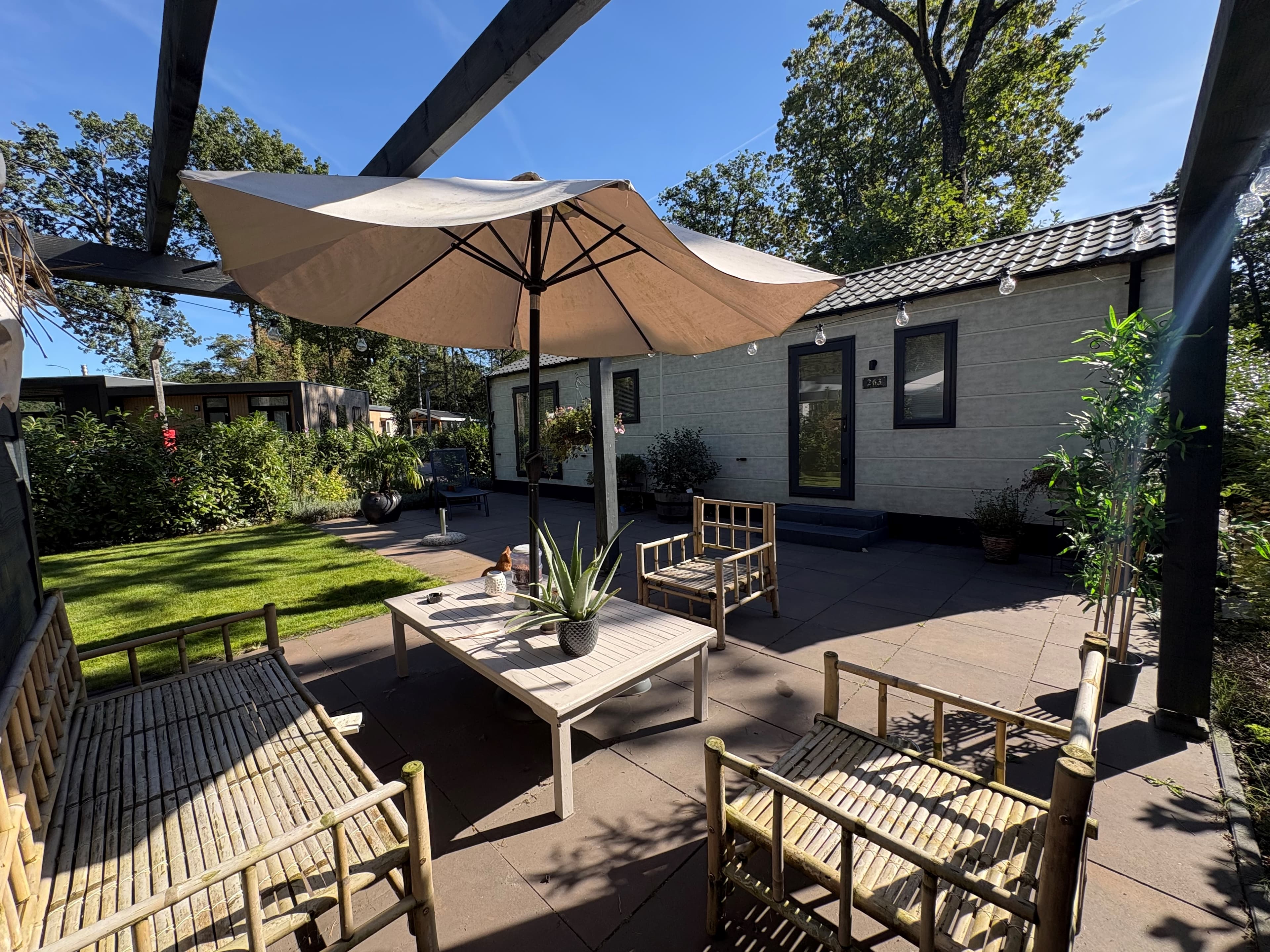 Terras met houten meubels en parasol voor een recreatiewoning omgeven door groen en een betegelde vloer