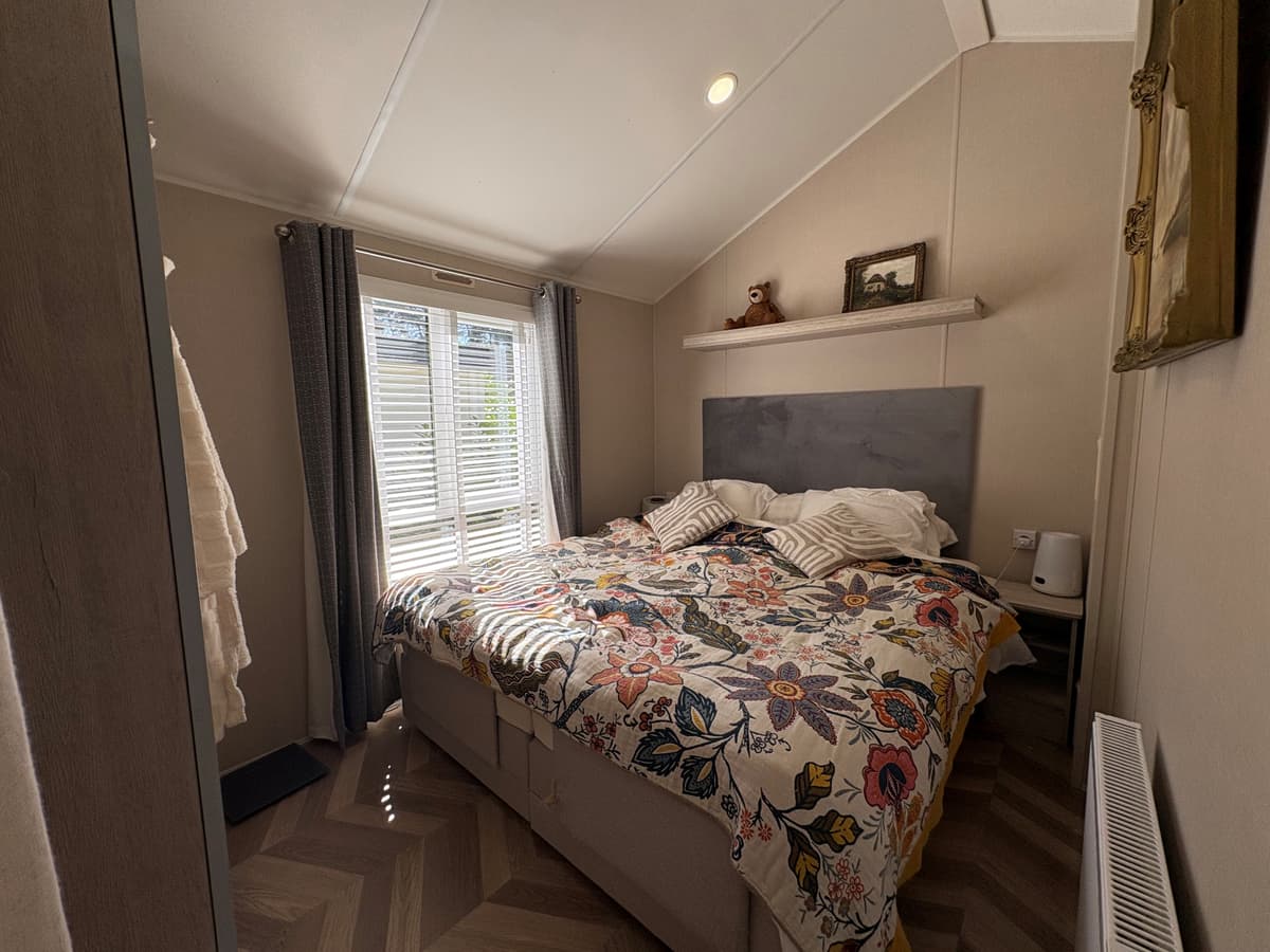 Slaapkamer met een tweepersoonsbed, kleurrijk dekbedovertrek en lichtinval via houten jaloezieën.