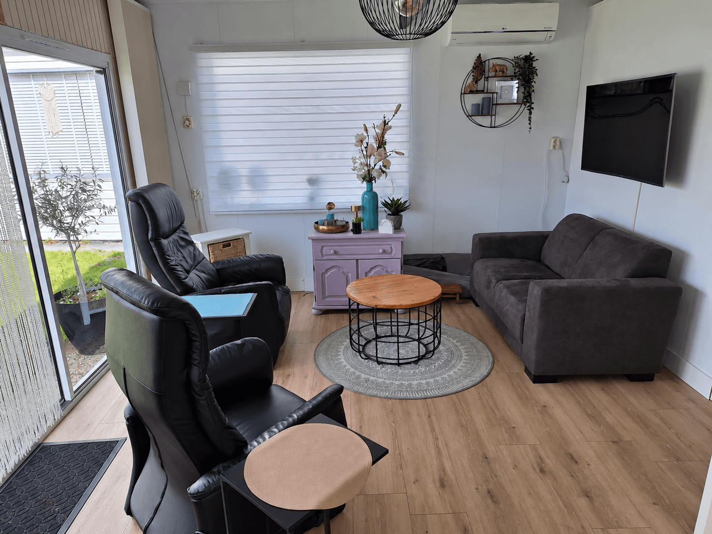 Woonkamer met zwarte fauteuils, bruine bank, ronde salontafel, en decoratie zoals bloemen en een tv aan de muur