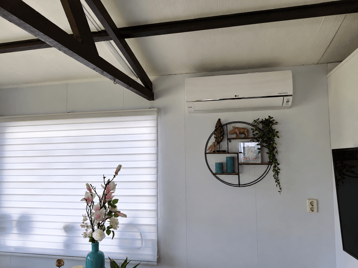Interieur van een recreatiewoning met wanddecoratie en airconditioning, enkele planten en een vaas met bloemen zichtbaar.