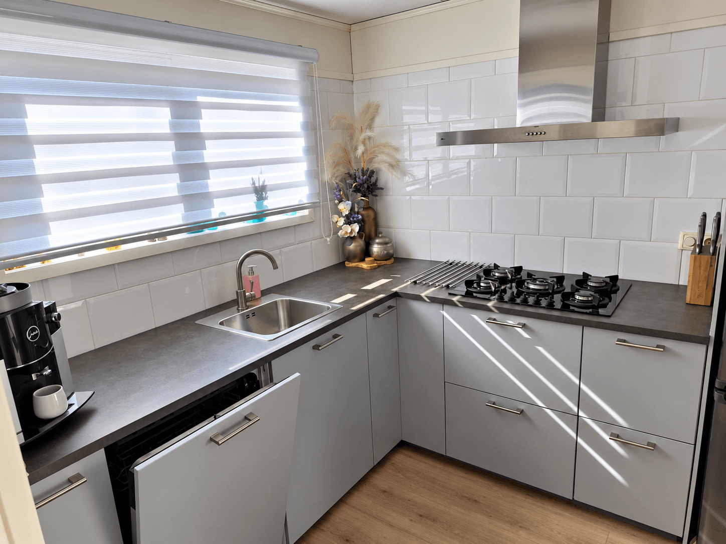 Moderne keuken met inbouwapparatuur, witte tegels, een spoelbak en een kookplaat, lichtinval door jaloezieën.