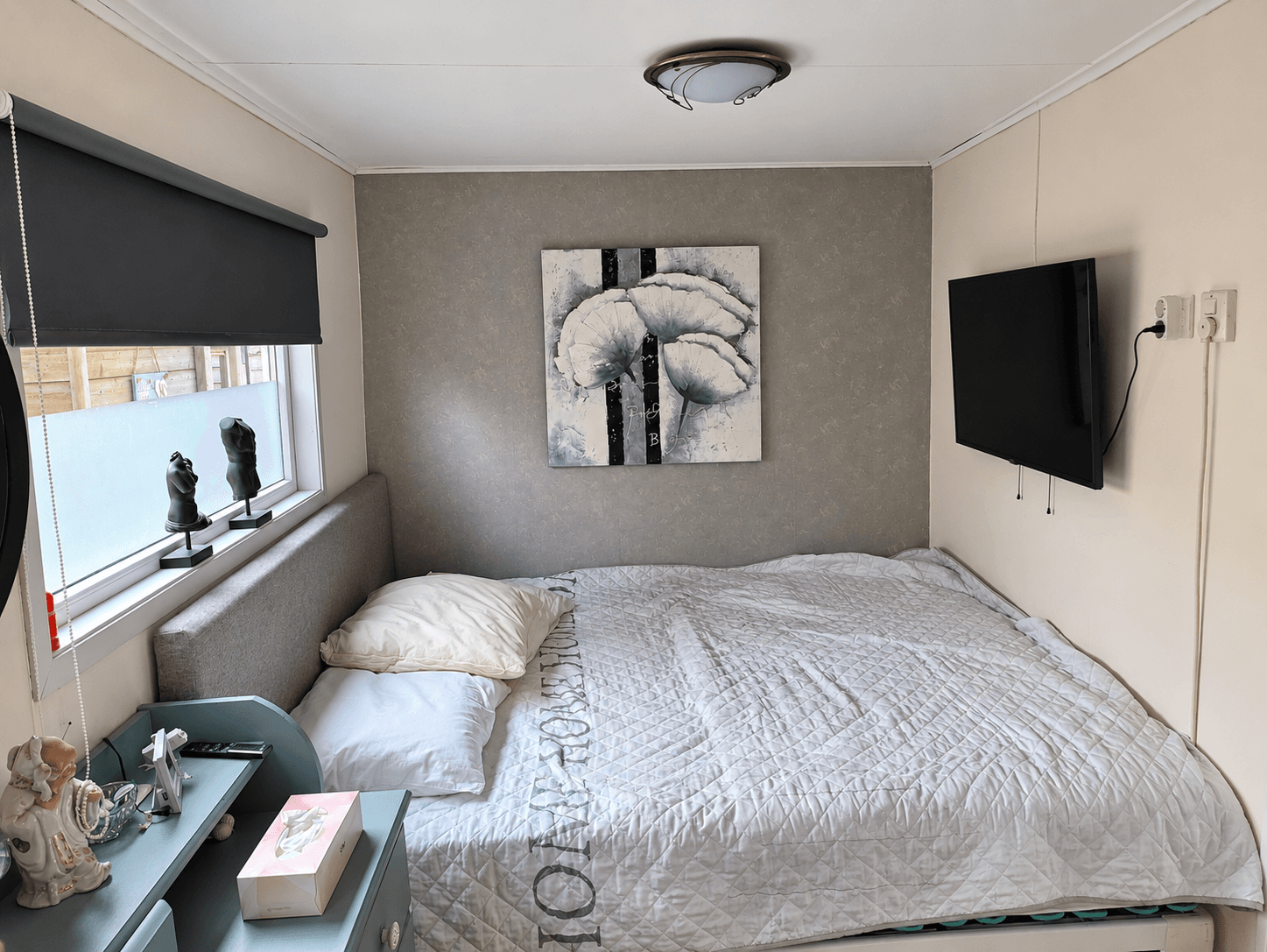 Slaapkamer met een tweepersoonsbed, schilderij aan de muur, tv en venster met zonwering en decoratieve beelden.