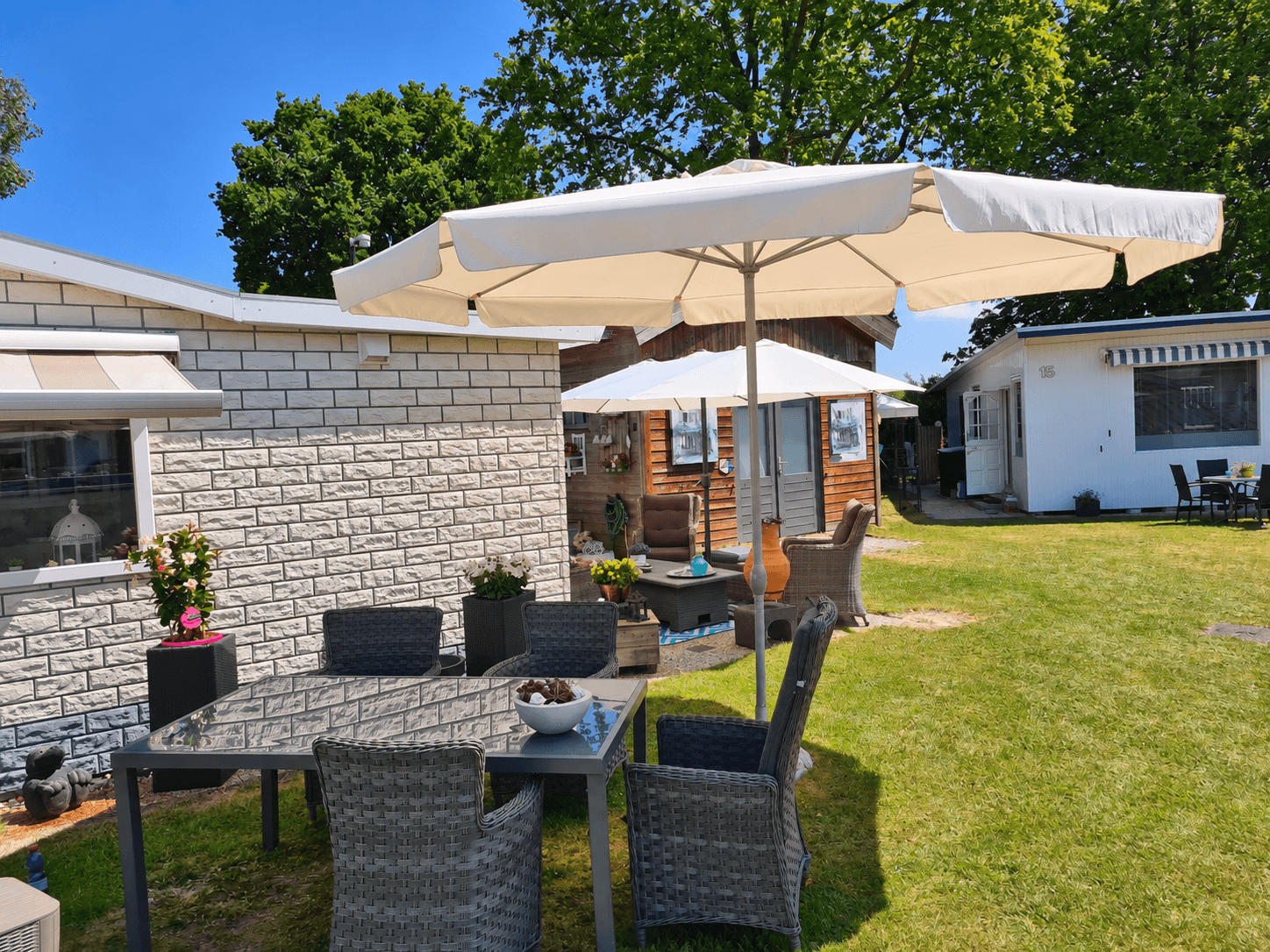 Tuin met een terras en een grote parasol, omringd door recreatiewoningen en groene bomen.
