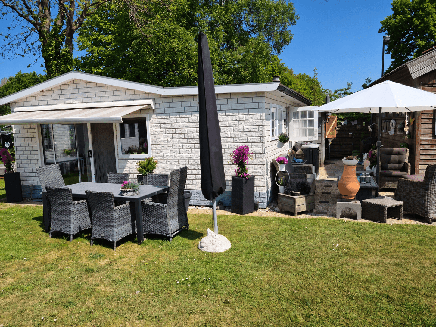 Recreatiewoning met terrasmeubelen, parasol en plantenbakken, omgeven door gras en bomen.