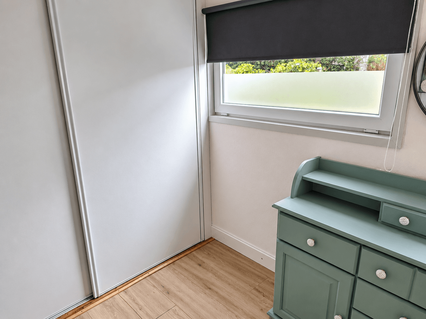Hoek van een lichte kamer met een raam, een grijze rolgordijn en een groene kast met lades