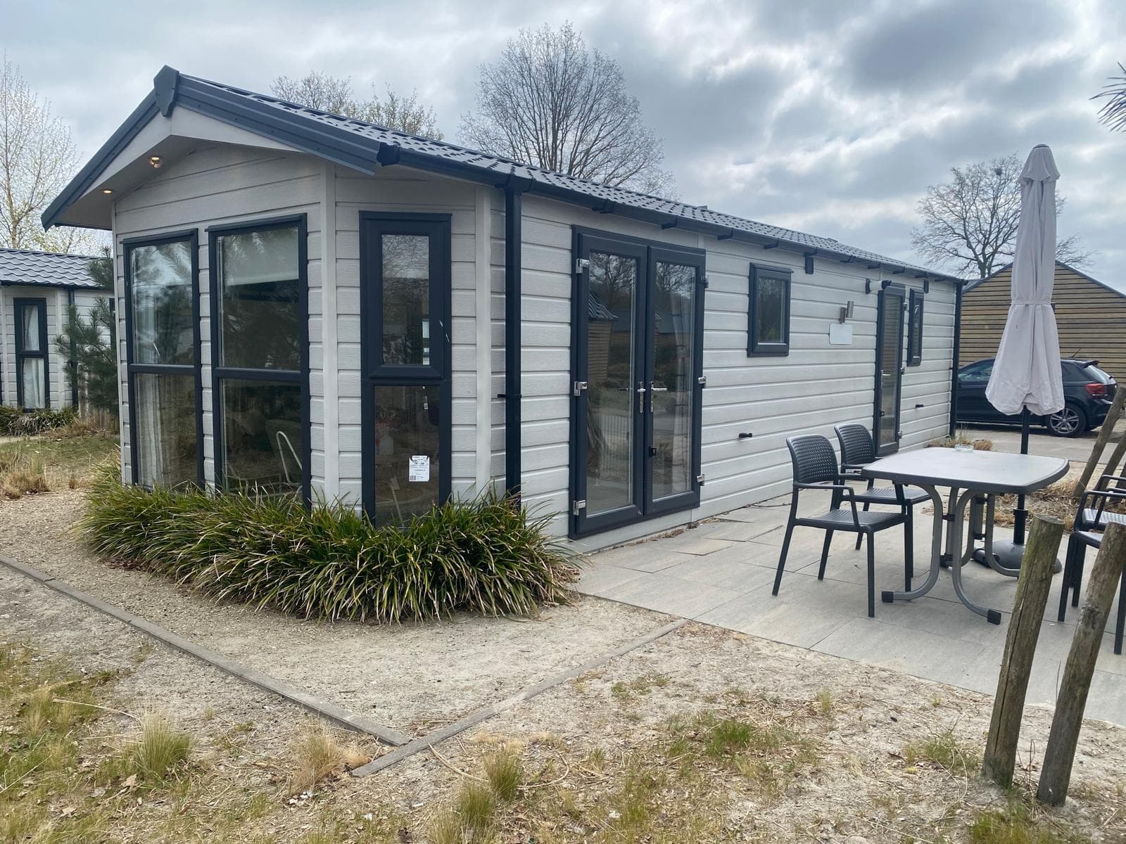Moderne vakantiewoning met grote ramen en een terras, omgeven door groen en zandgrond.