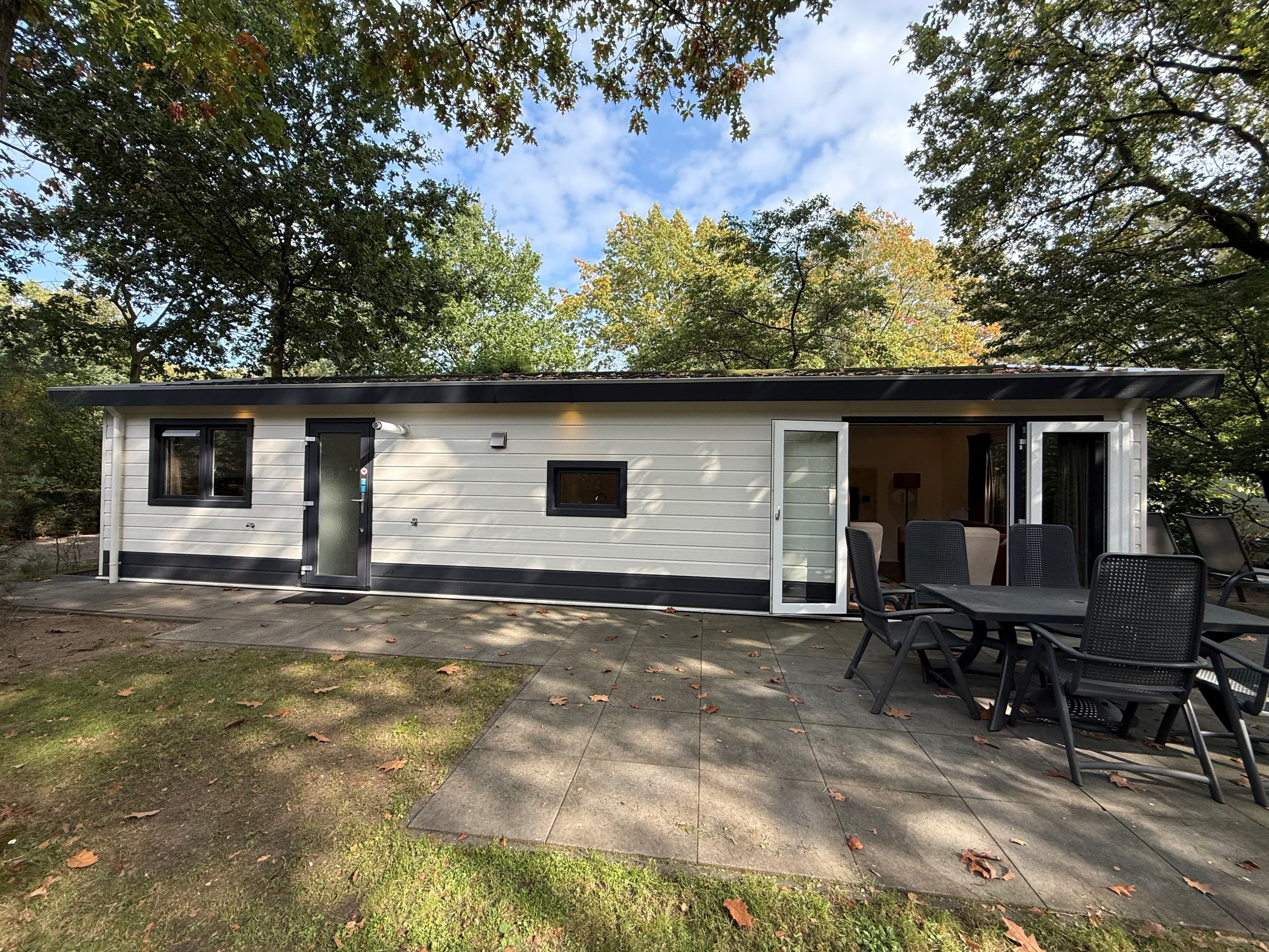 Moderne recreatiewoning met houten gevel, terras en tuinmeubilair, omgeven door bomen en groen.