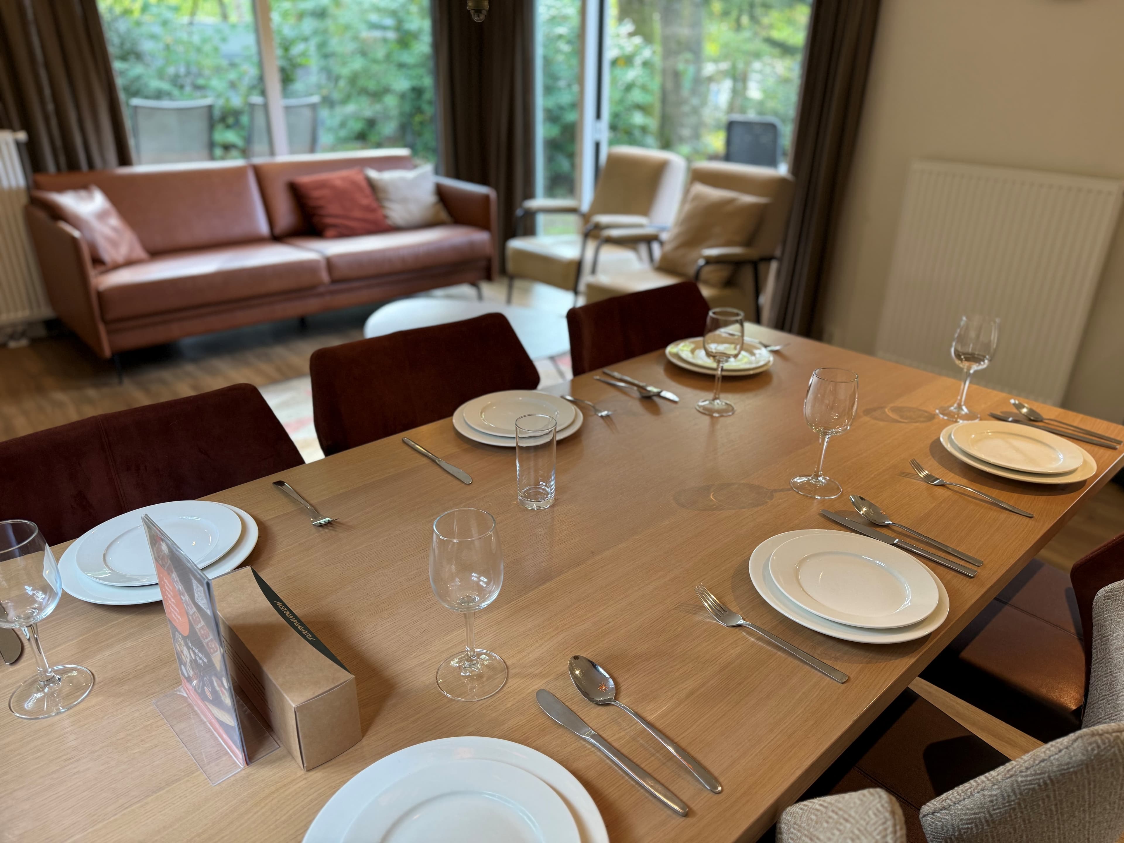 Eetkamertafel met servies en glazen, zicht op zithoek met een bank en grote ramen naar de tuin