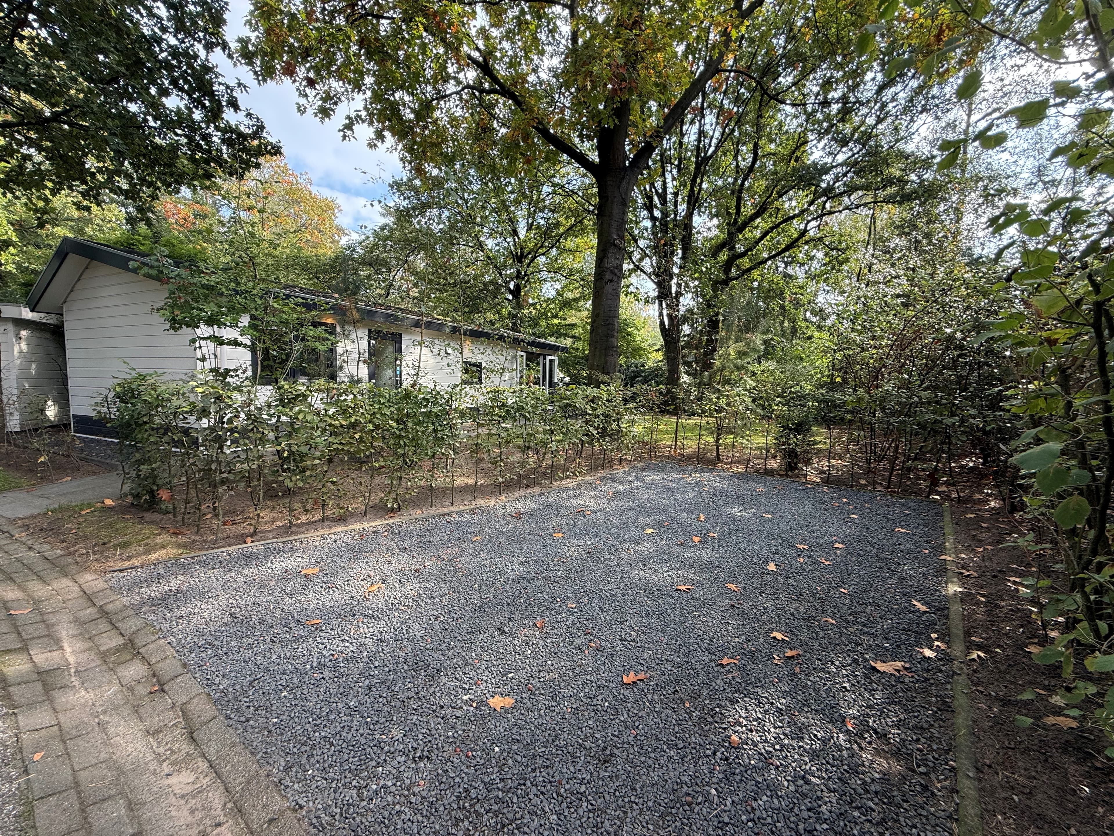 Grindplek voor parkeren omgeven door struiken en bomen met een vakantiewoning op de achtergrond.