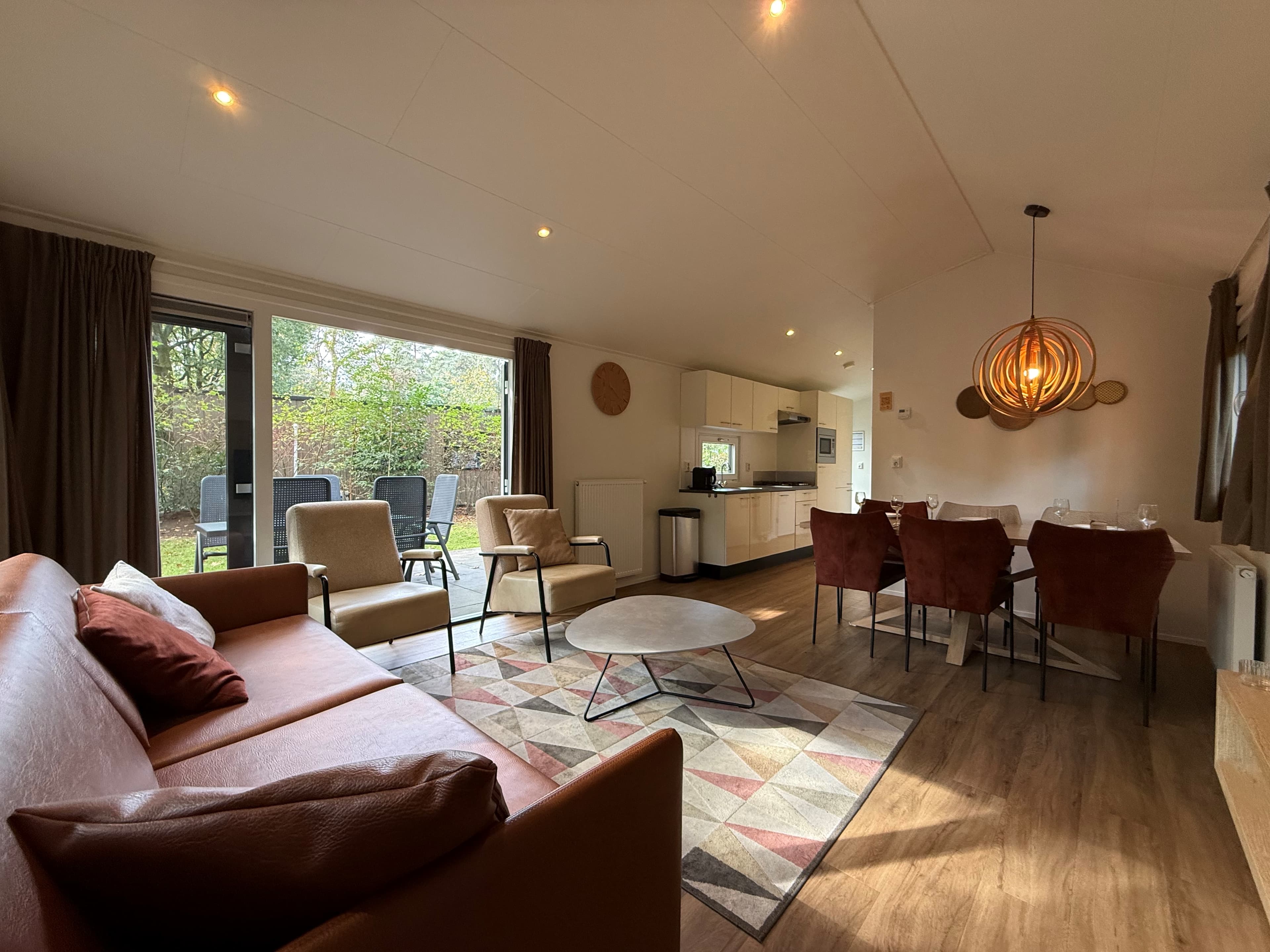 Moderne woonkamer met open keuken, grote ramen en toegang tot een terras met tafels en stoelen aan de achterzijde.