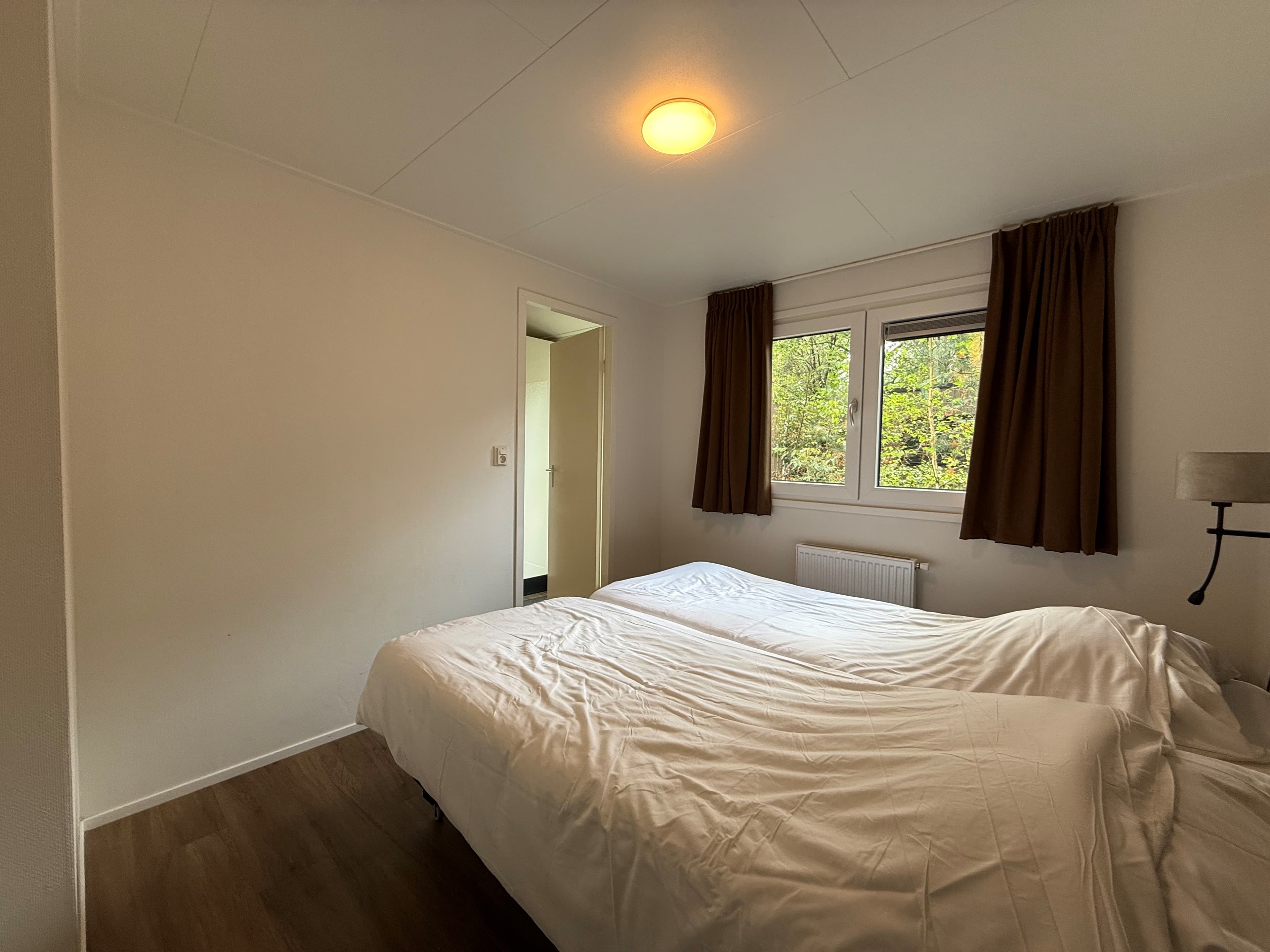 Slaapkamer met twee eenpersoonsbedden, lichtgekleurde muren en uitzicht op het groen door twee ramen met gordijnen