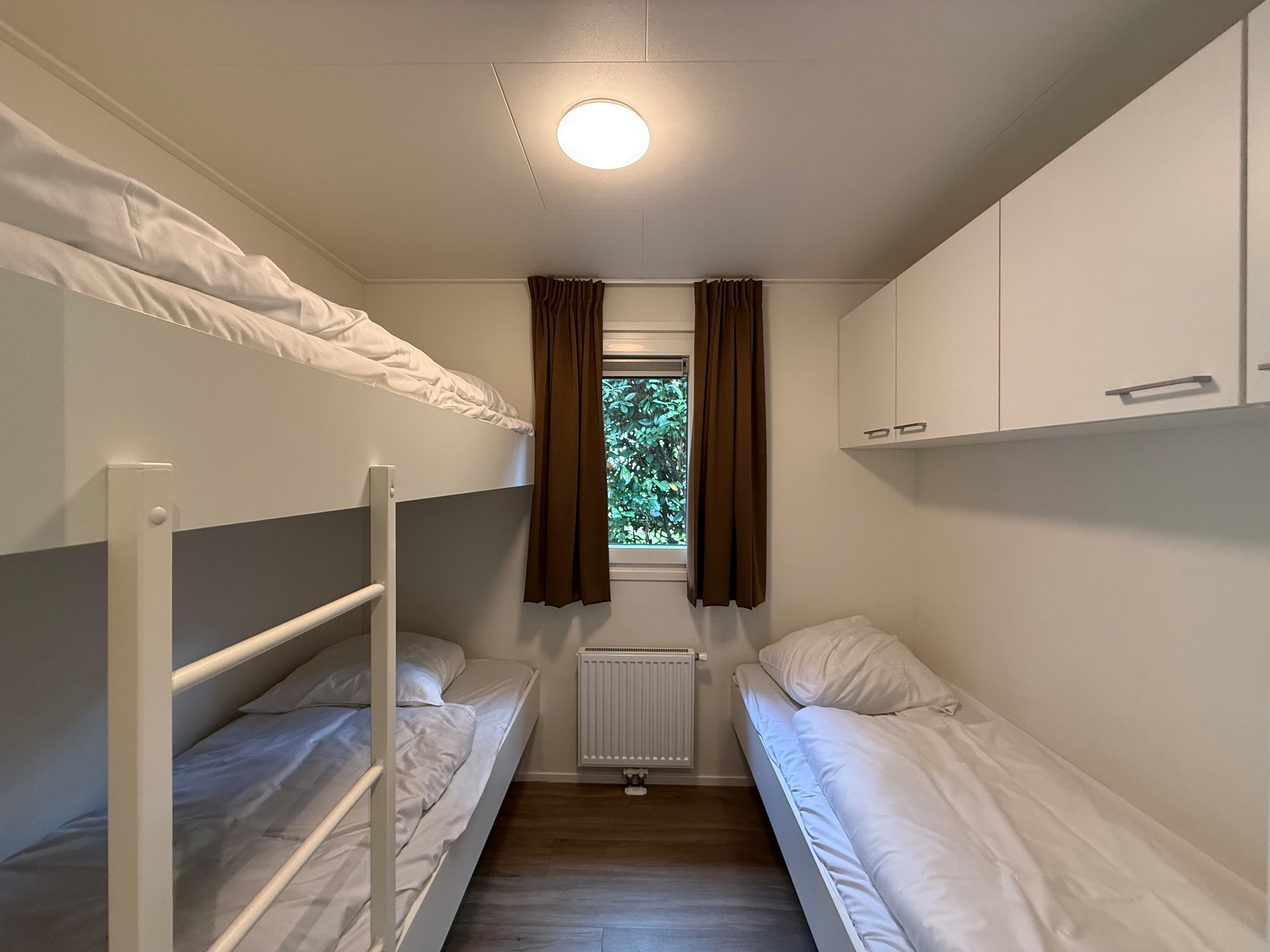 Slaapkamer met stapelbed en eenpersoonsbed, witte kasten en raam met uitzicht op begroeiing
