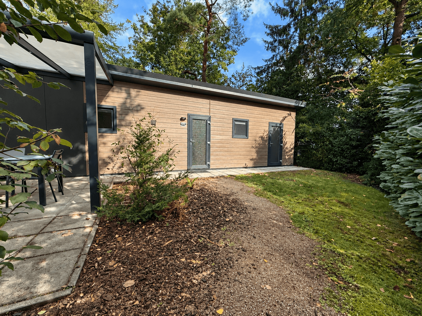 Recreatiewoning met houten gevel, tuinpad en omliggende bomen en struiken op Veluwepark de Bosgraaf in Lieren