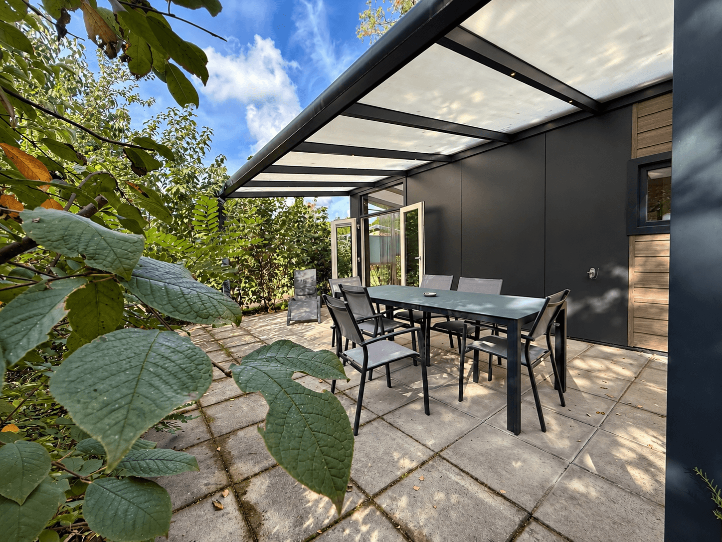 Terras met een eettafel en stoelen, omgeven door groene planten en een grijze stenen wand, onder een schuifdak