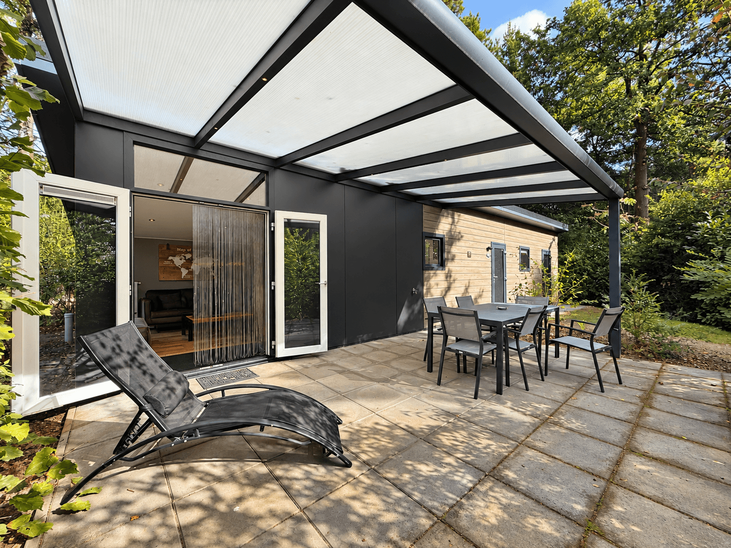 Terras met zithoek en ligstoel, overdekt met een doorzichtige bekleding, aan een recreatiewoning omringd door groen