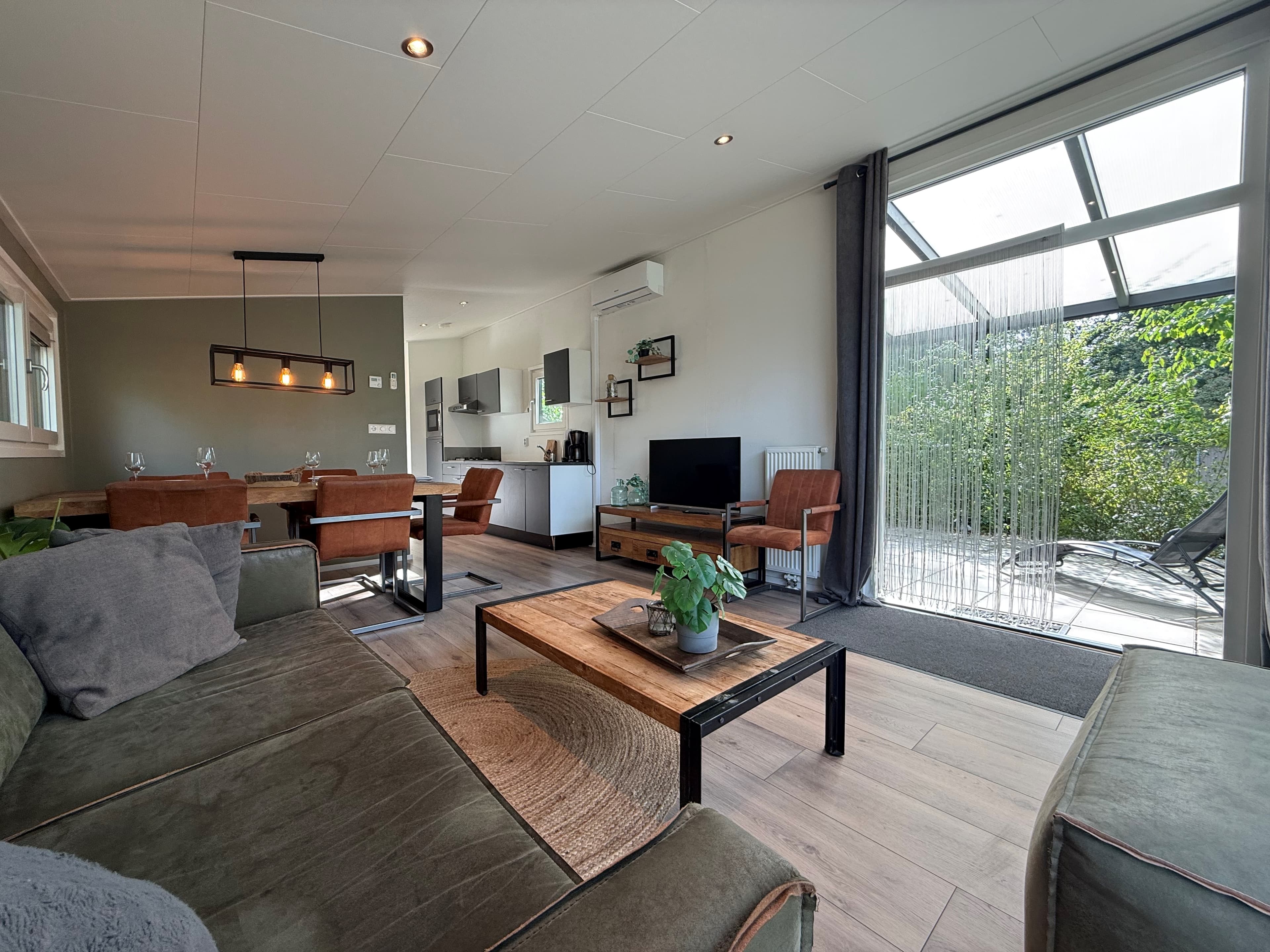 Gezellige woonkamer met bank, eethoek, keuken en een toegangsdeur naar de tuin met ramen en planten.