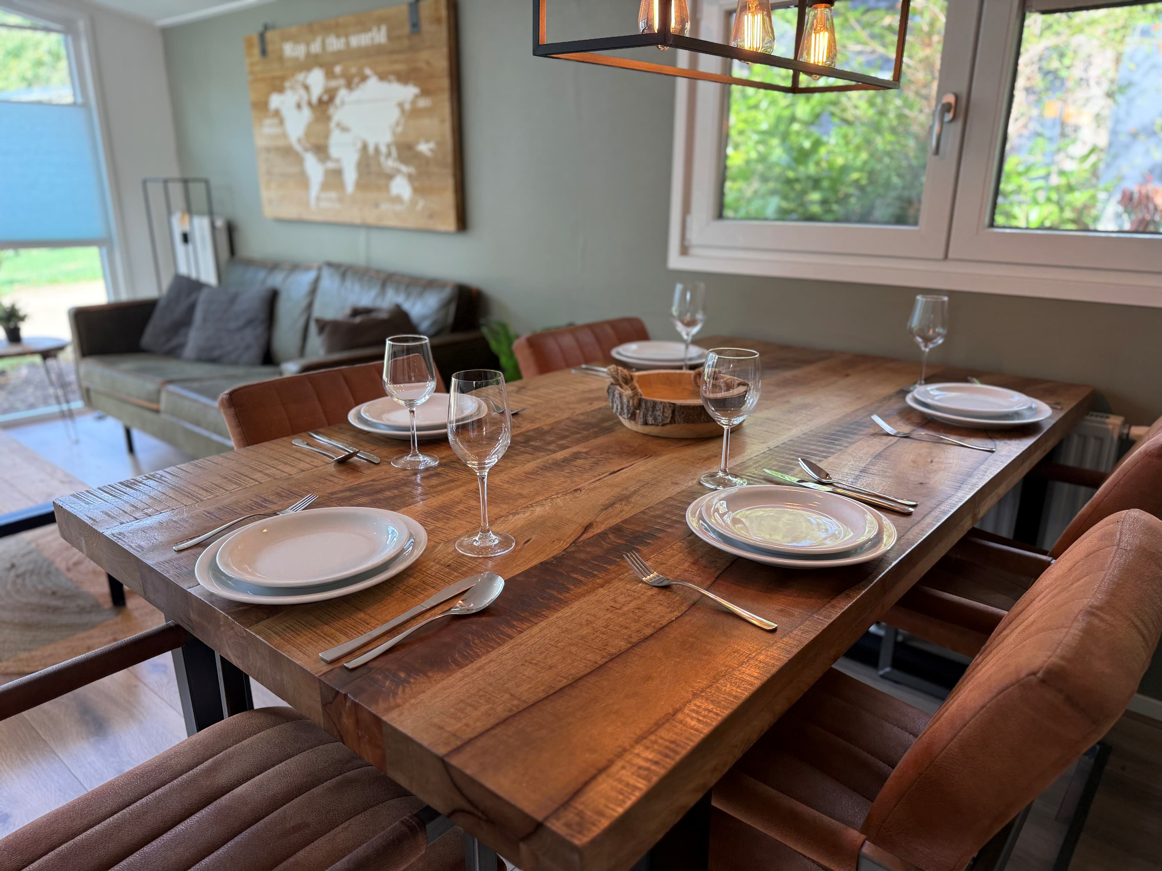 Houten eettafel met servies en glazen, moderne banken en een wereldkaart aan de muur in een sfeervolle woonkamer