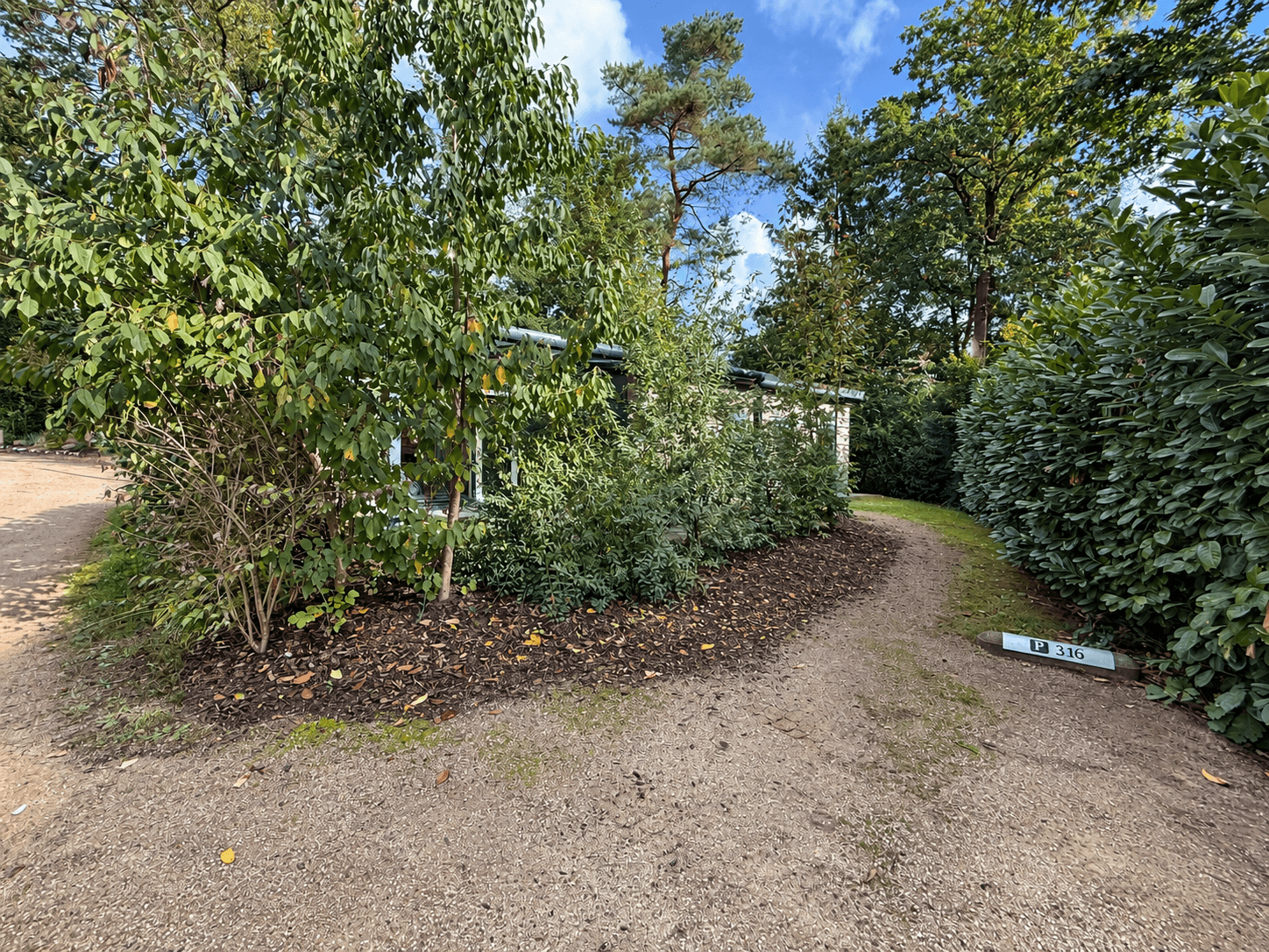 Pad omgeven door groen en bomen, met een recreatiewoning zichtbaar achter de struiken en een huisnummerbordje.