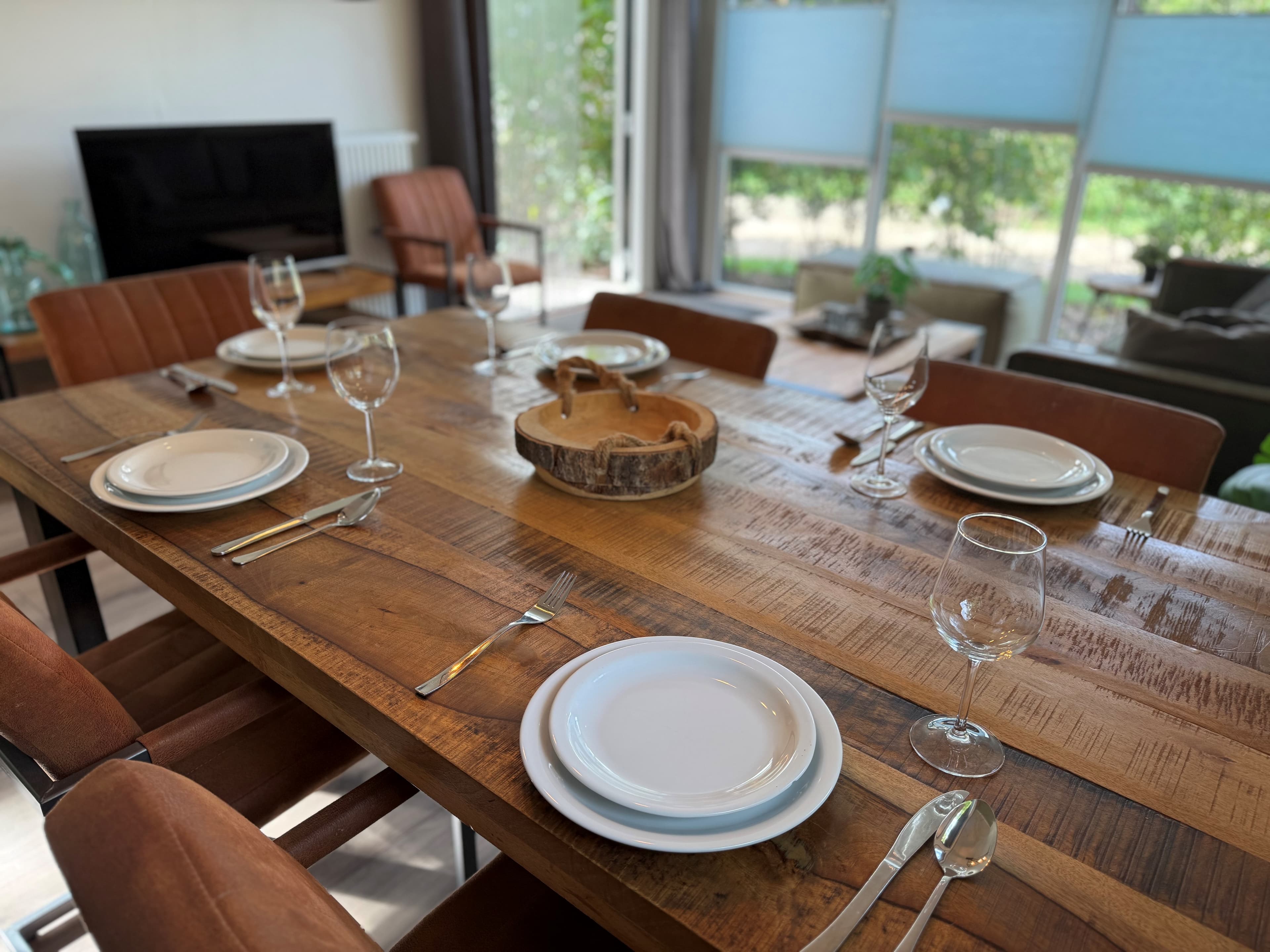 Eettafel met borden en glazen, omringd door stoelen, in een lichte woonkamer met zicht op de tuin