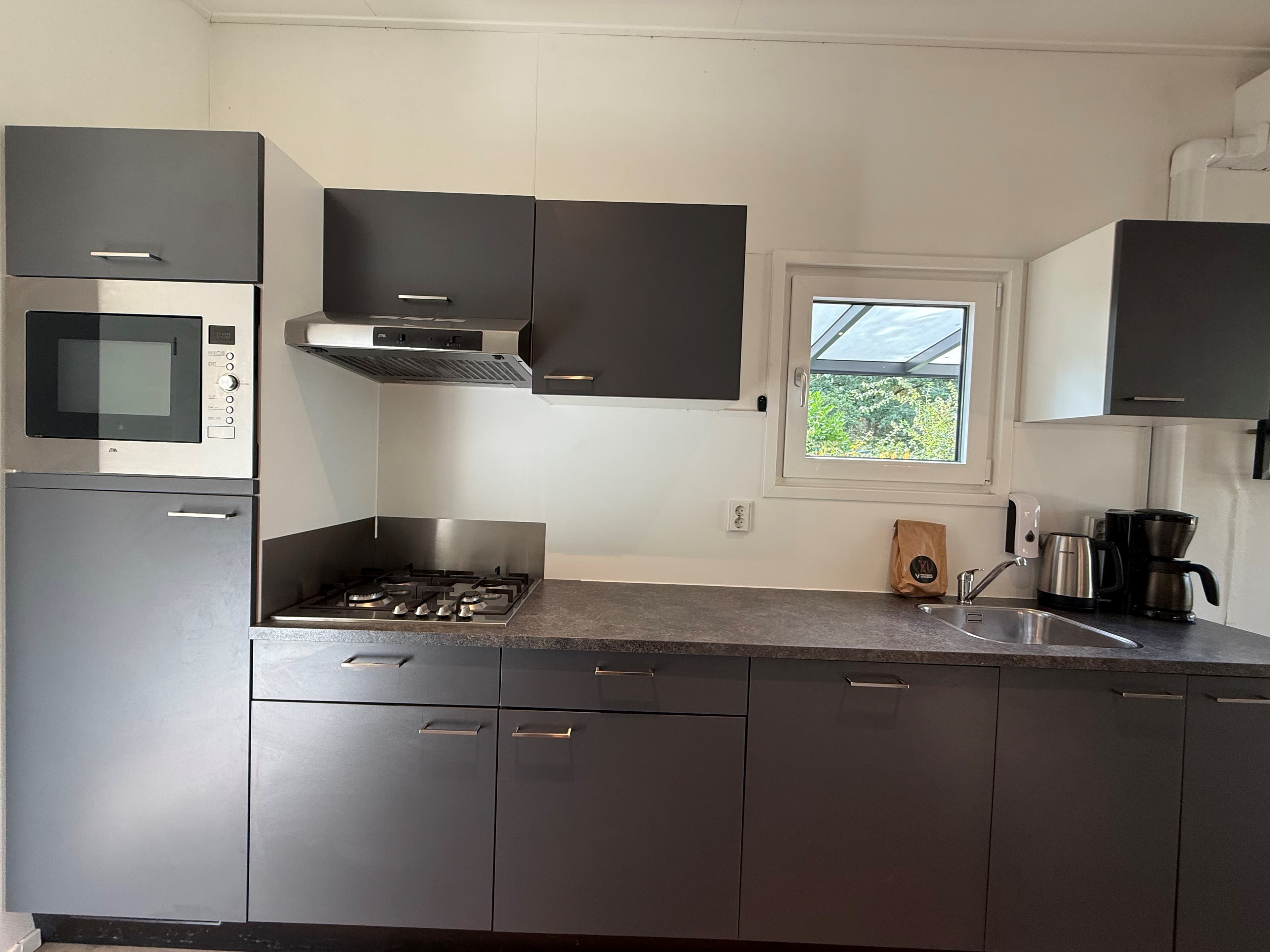 Moderne keuken met grijze kasten, gasfornuis, afzuigkap, oven, koelkast en koffiezetapparaat boven een werkblad