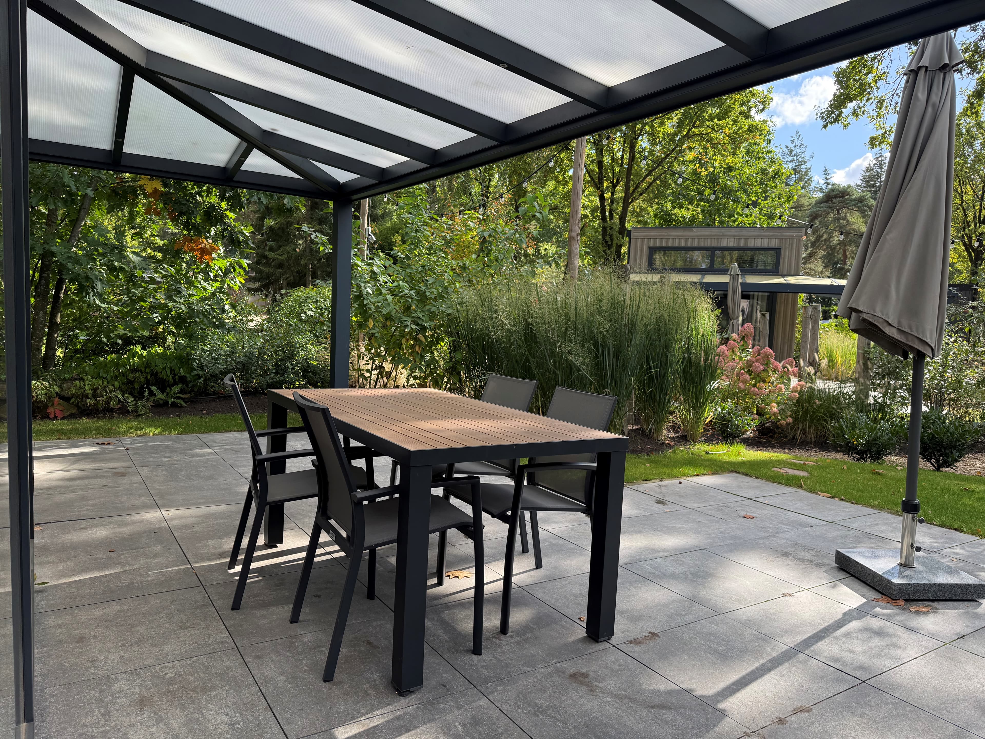 Tuinset met grote tafel en stoelen onder een overkapping, omgeven door groen en planten