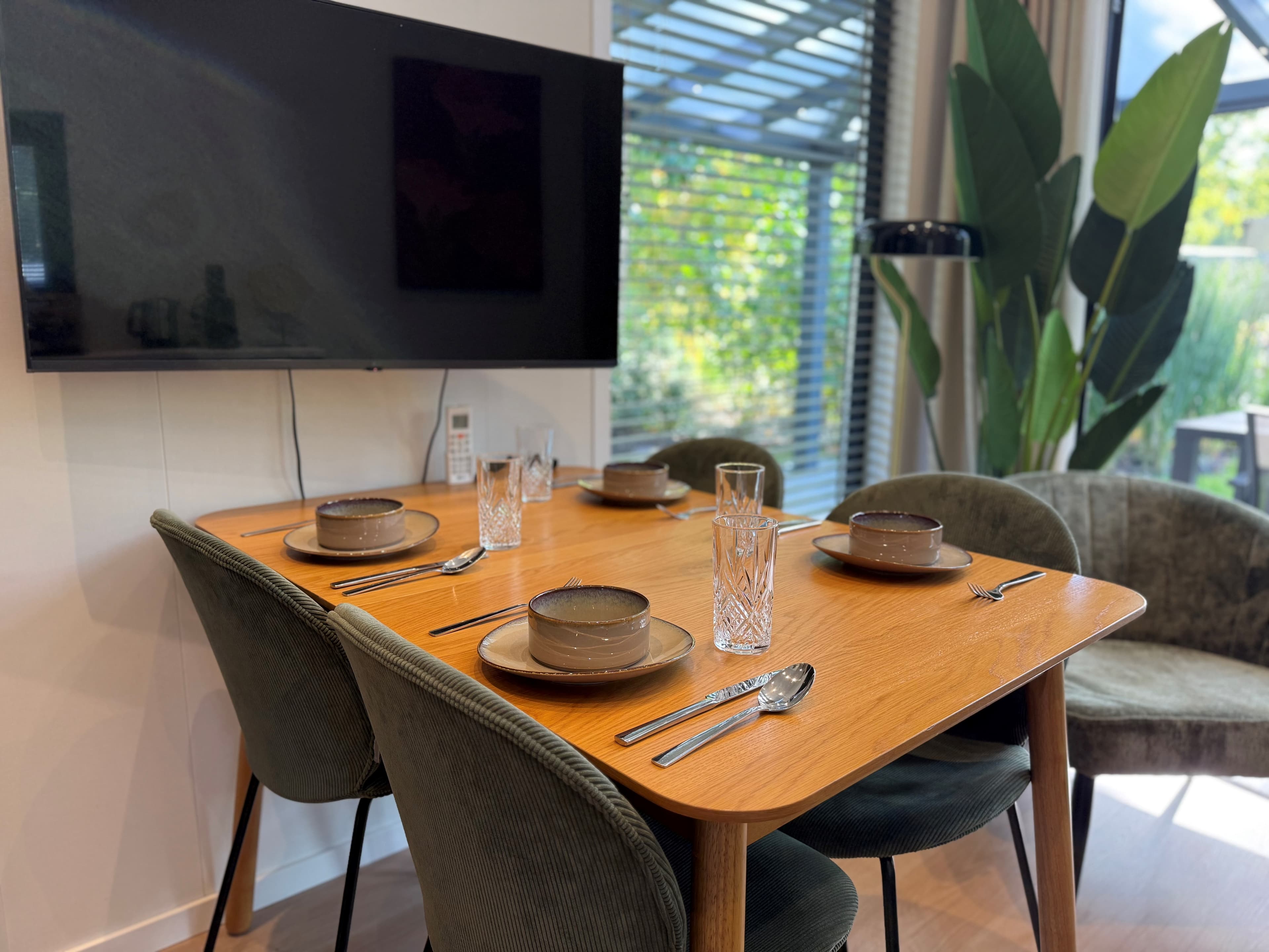 Eettafel met vier stoelen, servies en glazen, tegen een wand met een tv en uitzicht op groene buitenruimte.