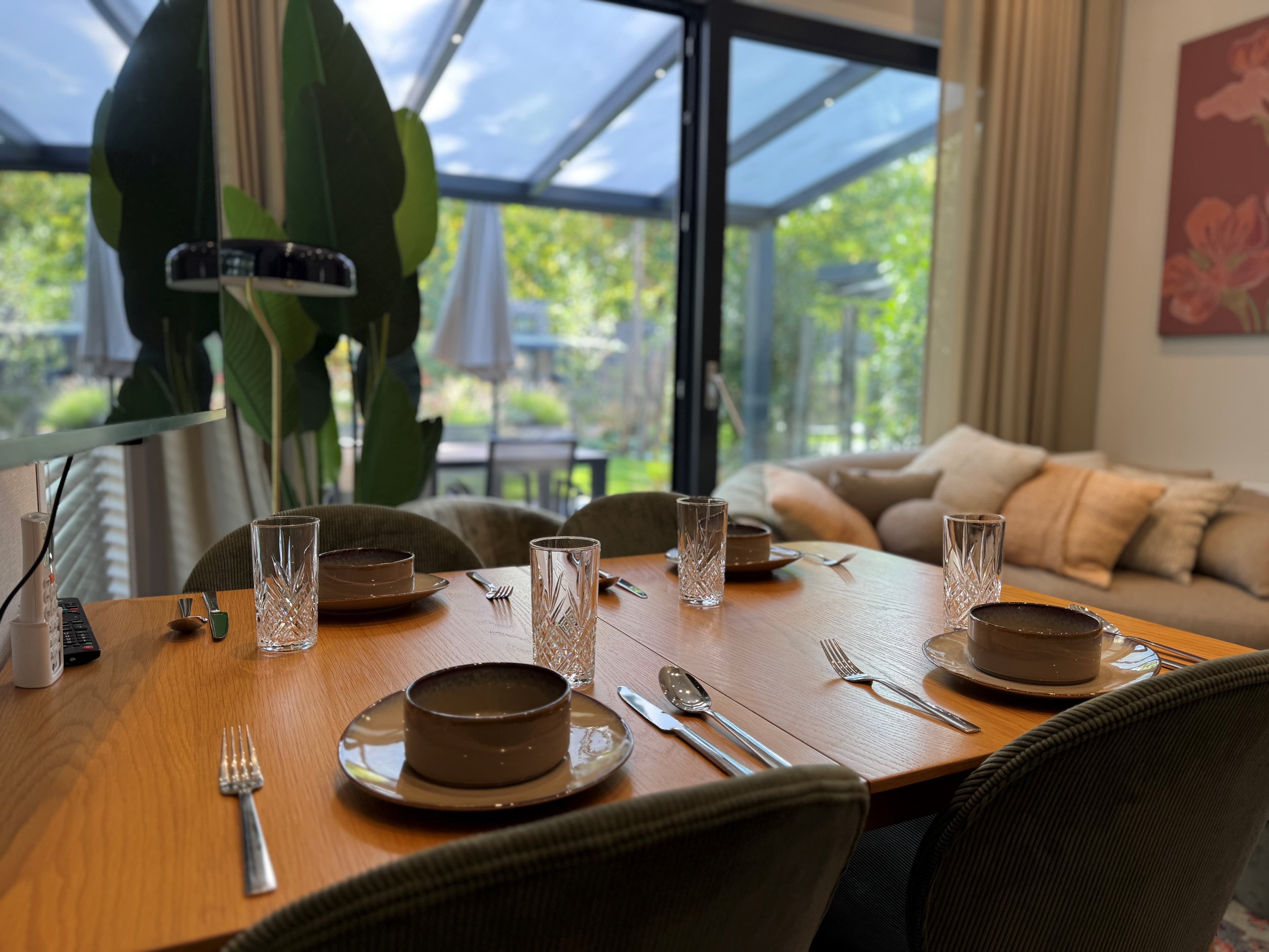 Eettafel met servies en glazen, zicht op binnenruimte met grote ramen en planten op de achtergrond