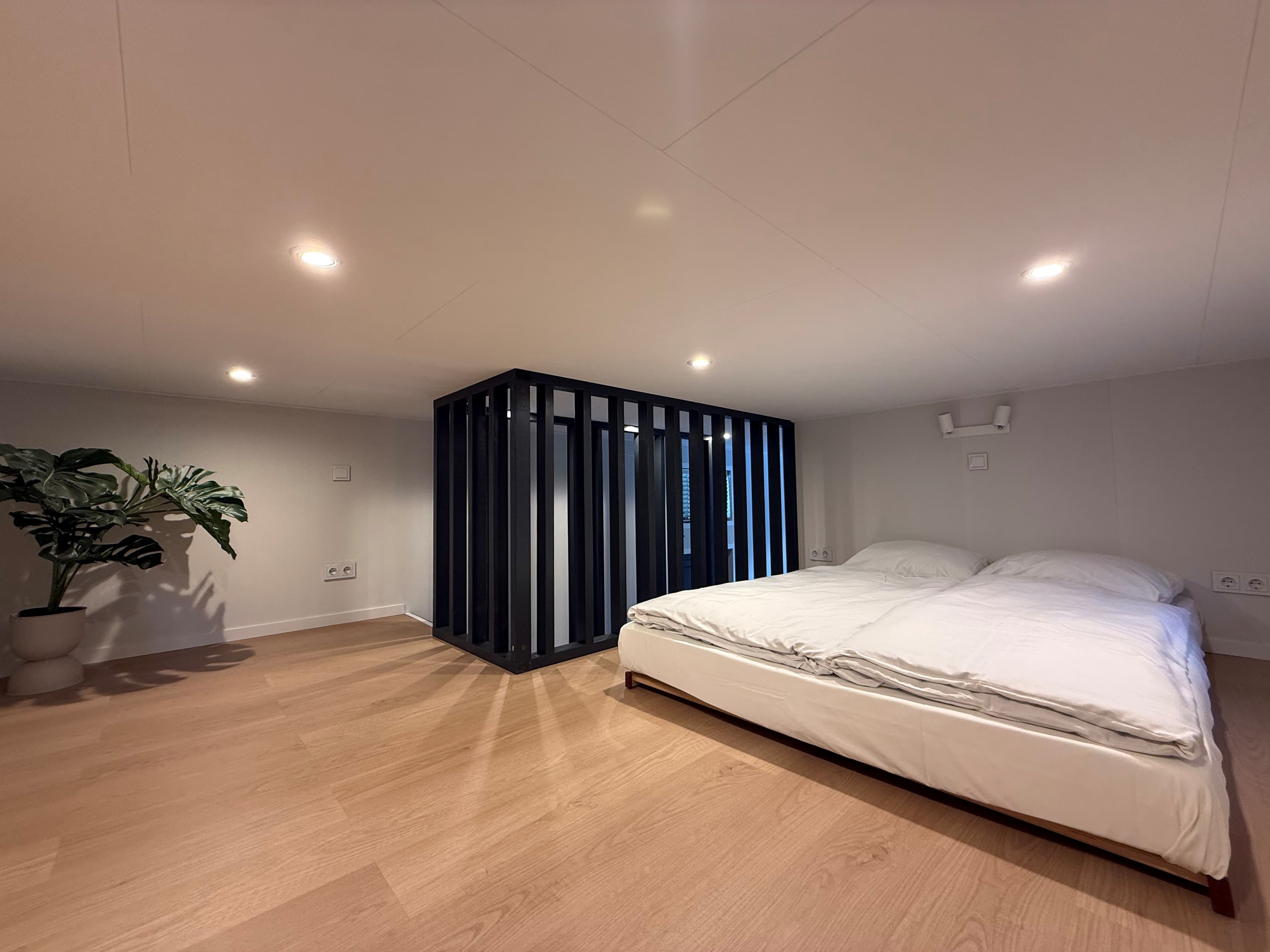 Slaapkamer met tweepersoonsbed, houten vloer en een groen plantje naast een zwarte afscheiding met lichtinval.