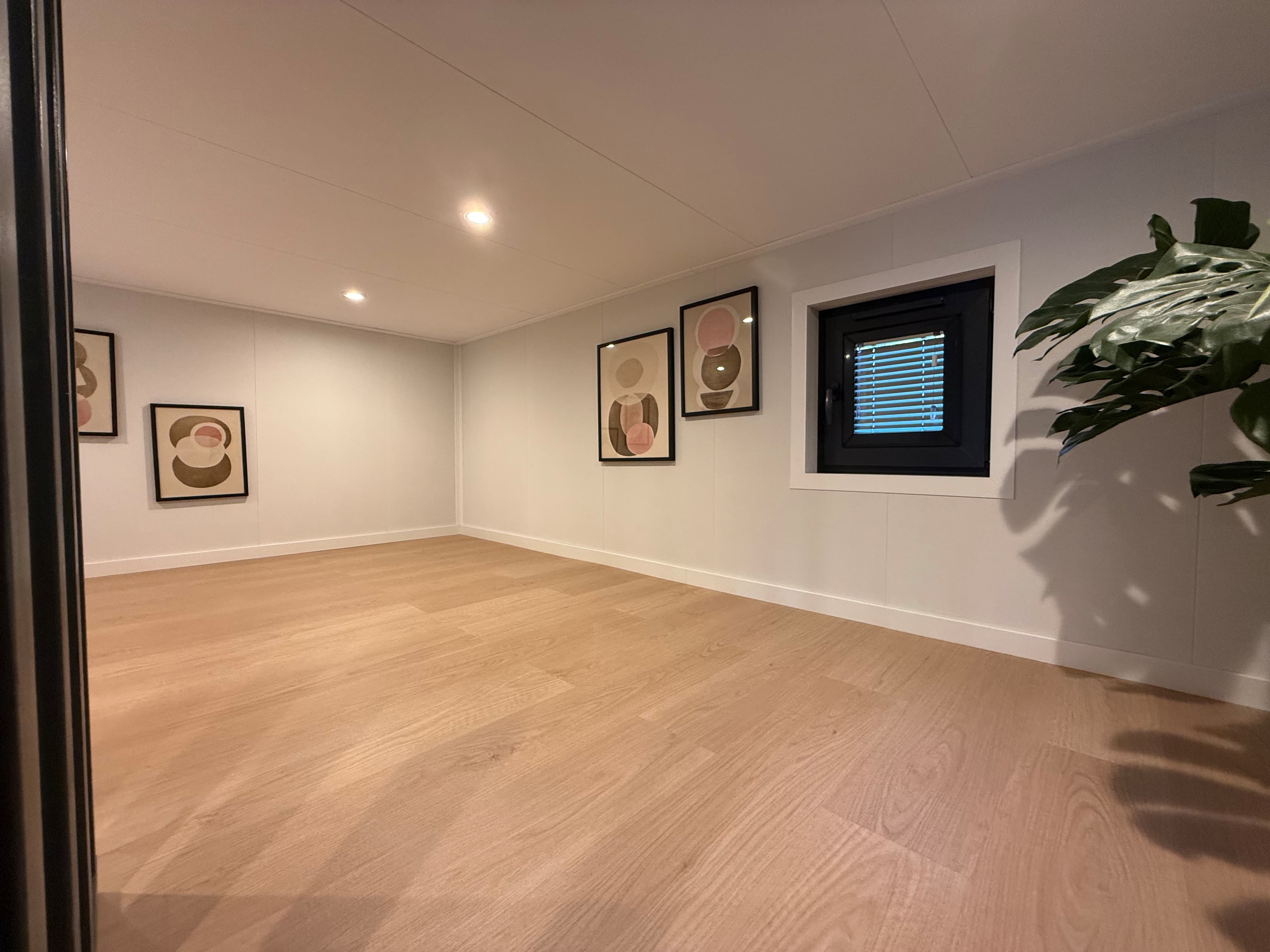 Ruime woonkamer met houten vloer, witte muren en kunst aan de muur, verlicht door inbouwverlichting.
