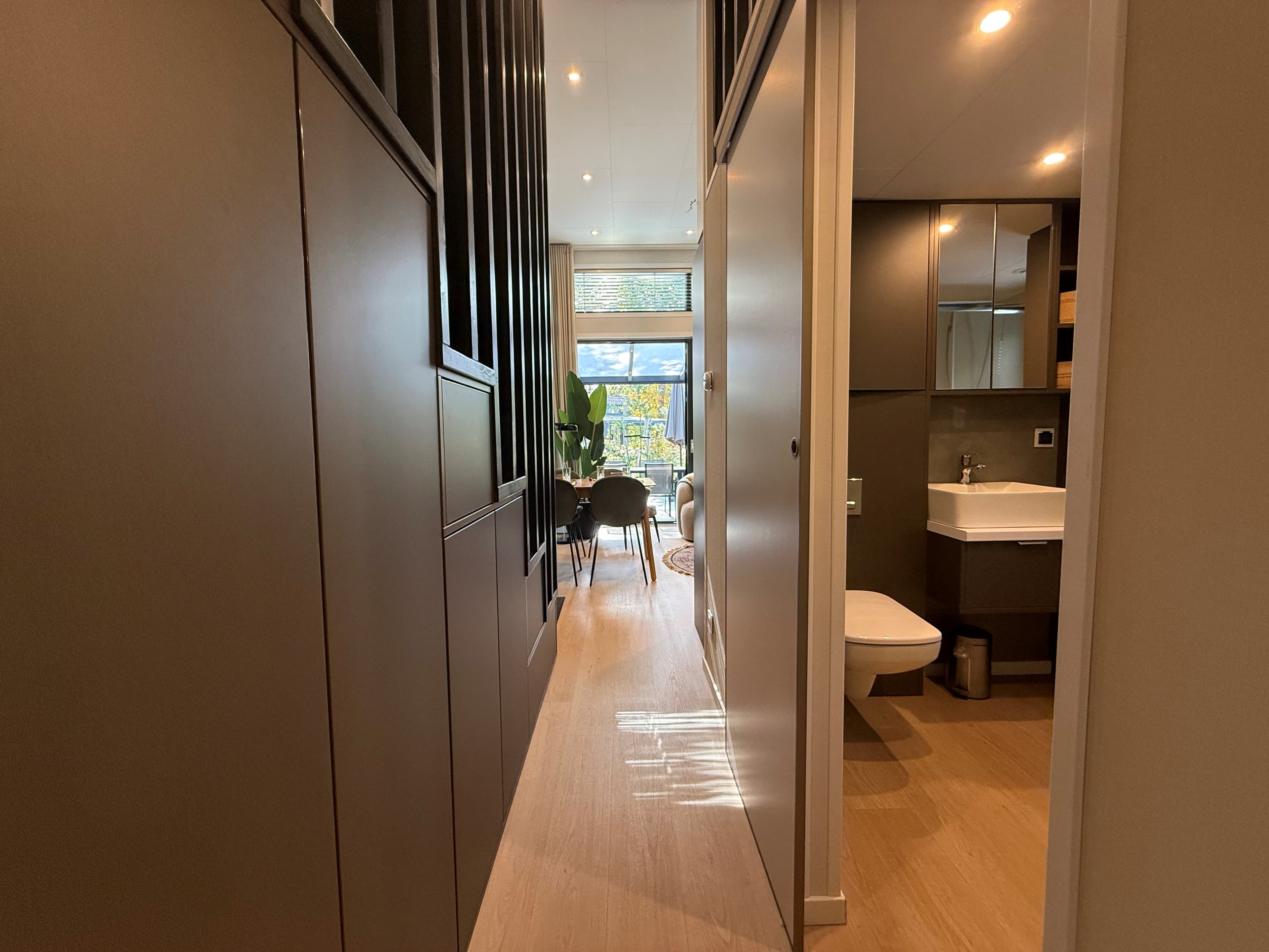 Lichte gang met toegang tot badkamer en woonkamer, met zicht op een eetgedeelte en raampartij.